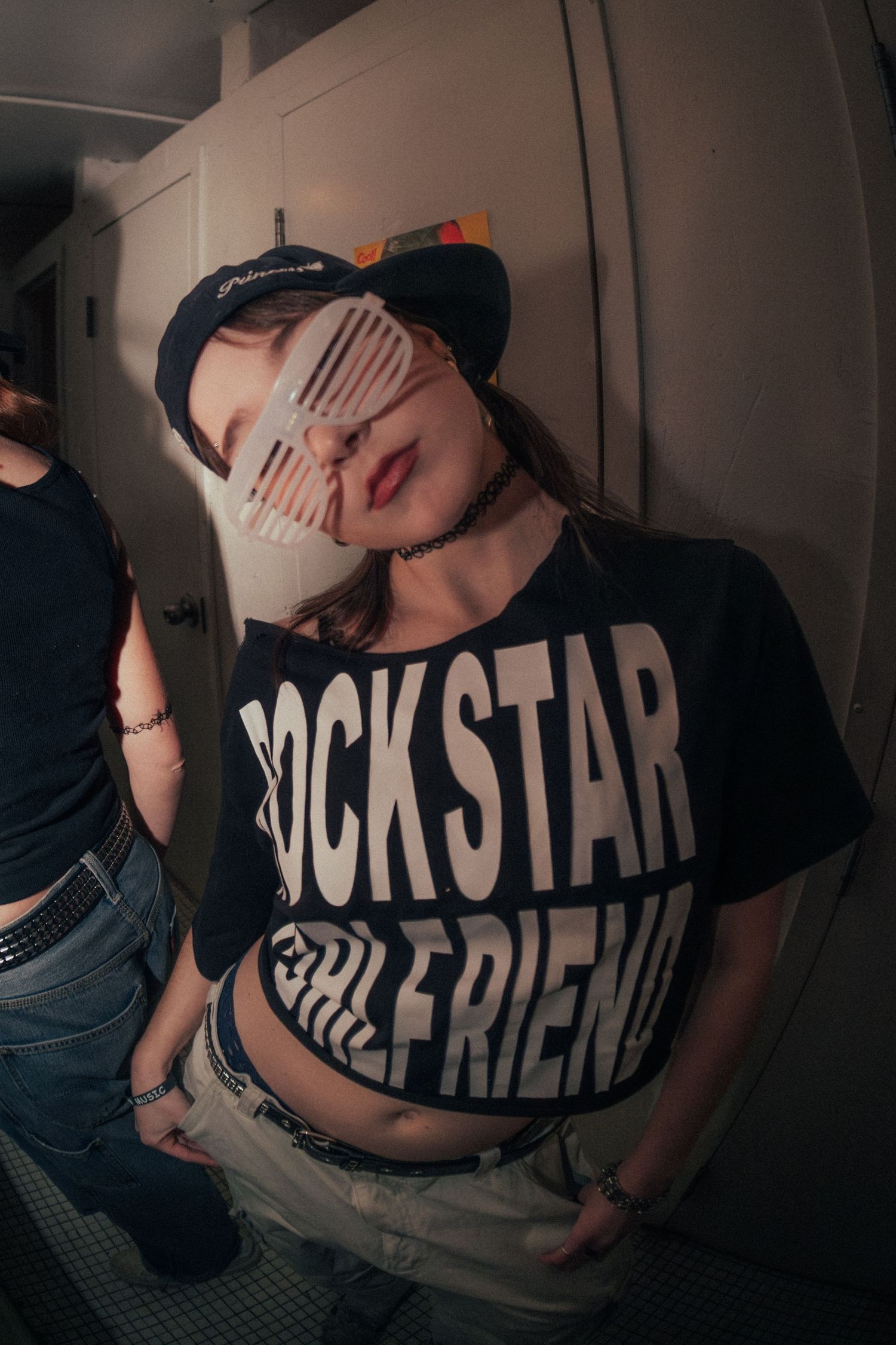 Young woman in a Rockstar Girlfriend crop tee, snapback cap, and shutter shades posing indoors.