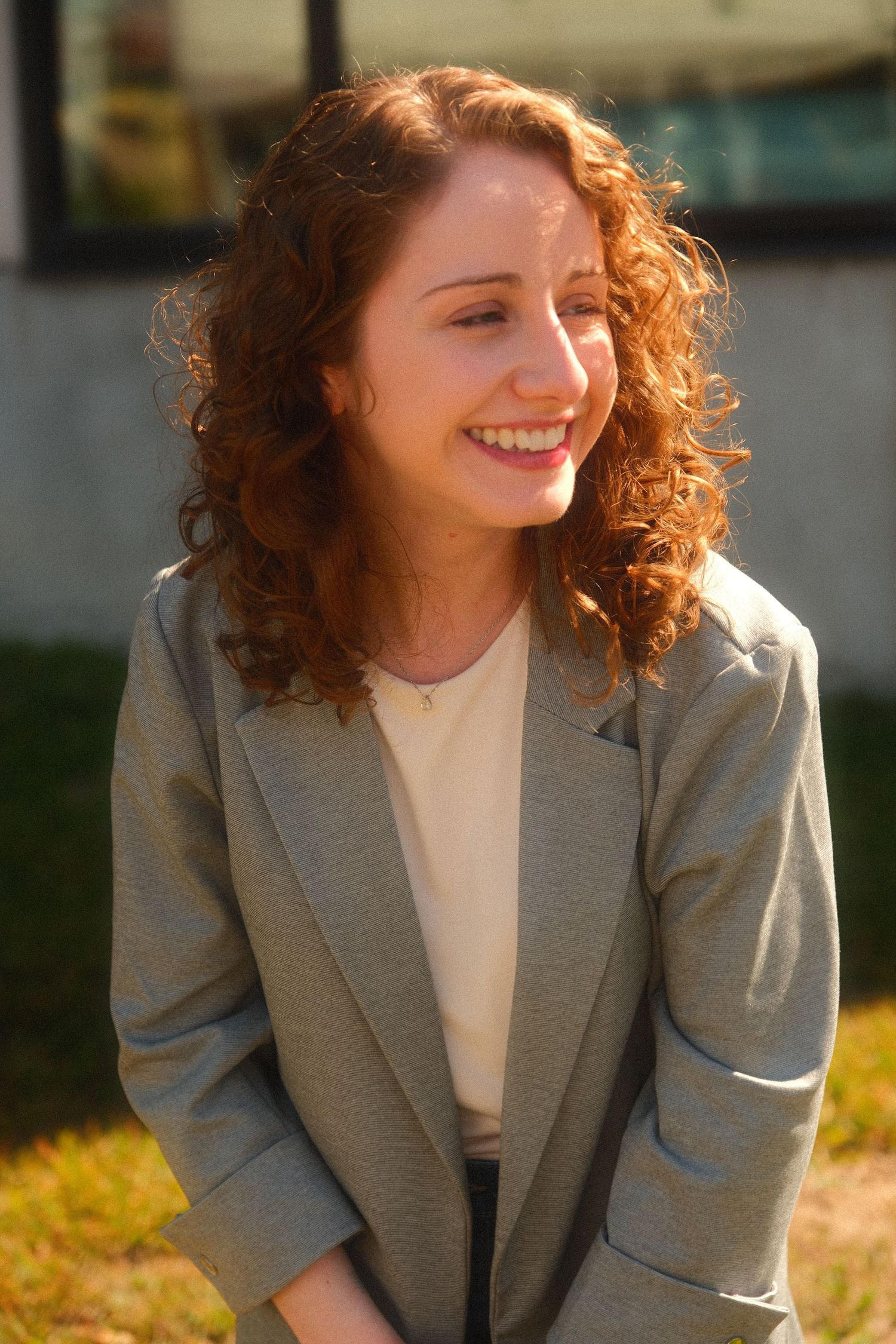 Young woman with curly red hair smiling outdoors in a gray blazer on a sunny day.