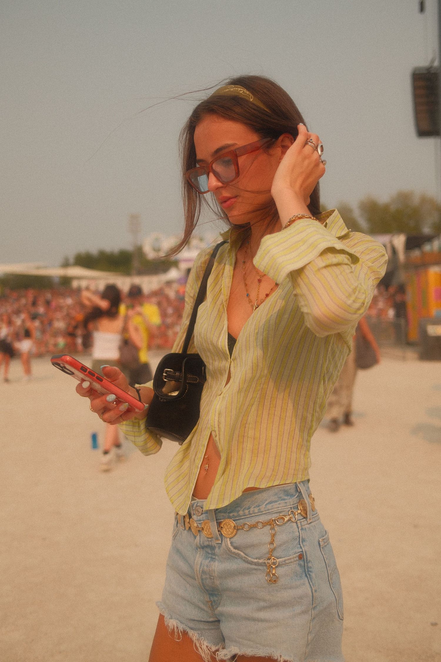 Young woman in yellow shirt, denim shorts, and sunglasses checks phone at outdoor music festival.
