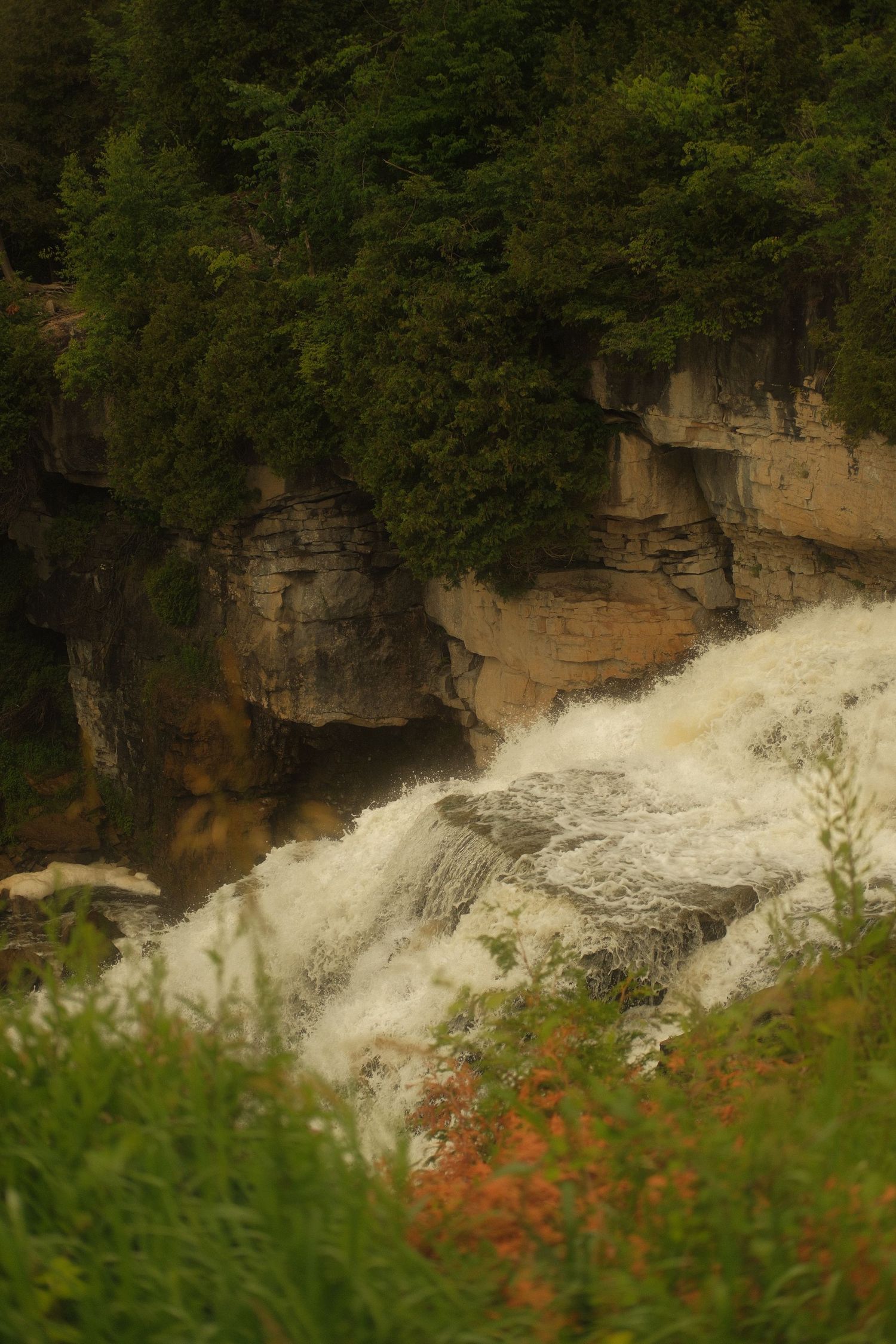 A powerful waterfall cascades over rugged limestone rocks surrounded by dense green forest vegetation.