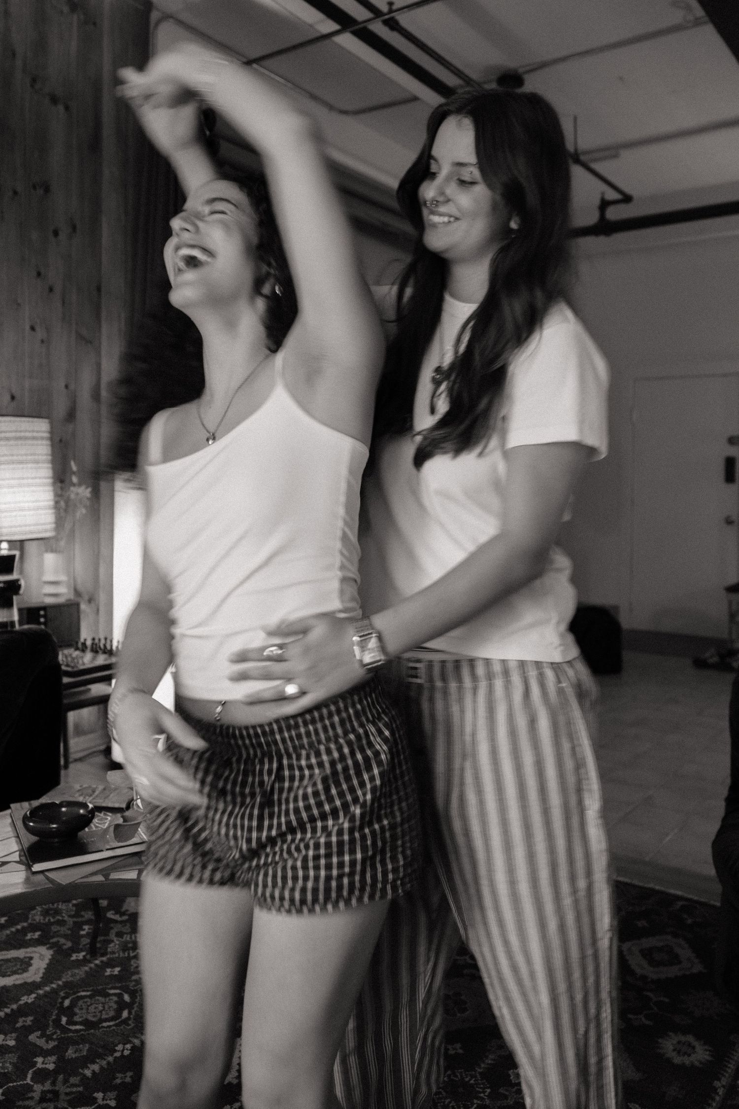 Two women laugh and dance together in a black and white photo indoors.