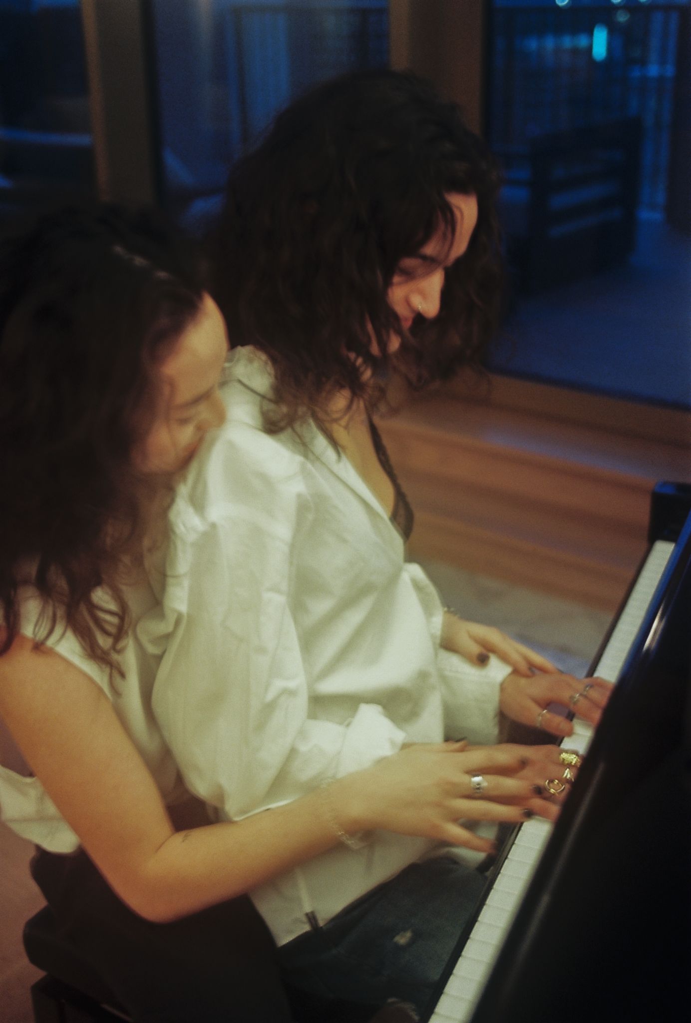 Two women with curly hair play piano together, smiling warmly in a cozy evening setting.