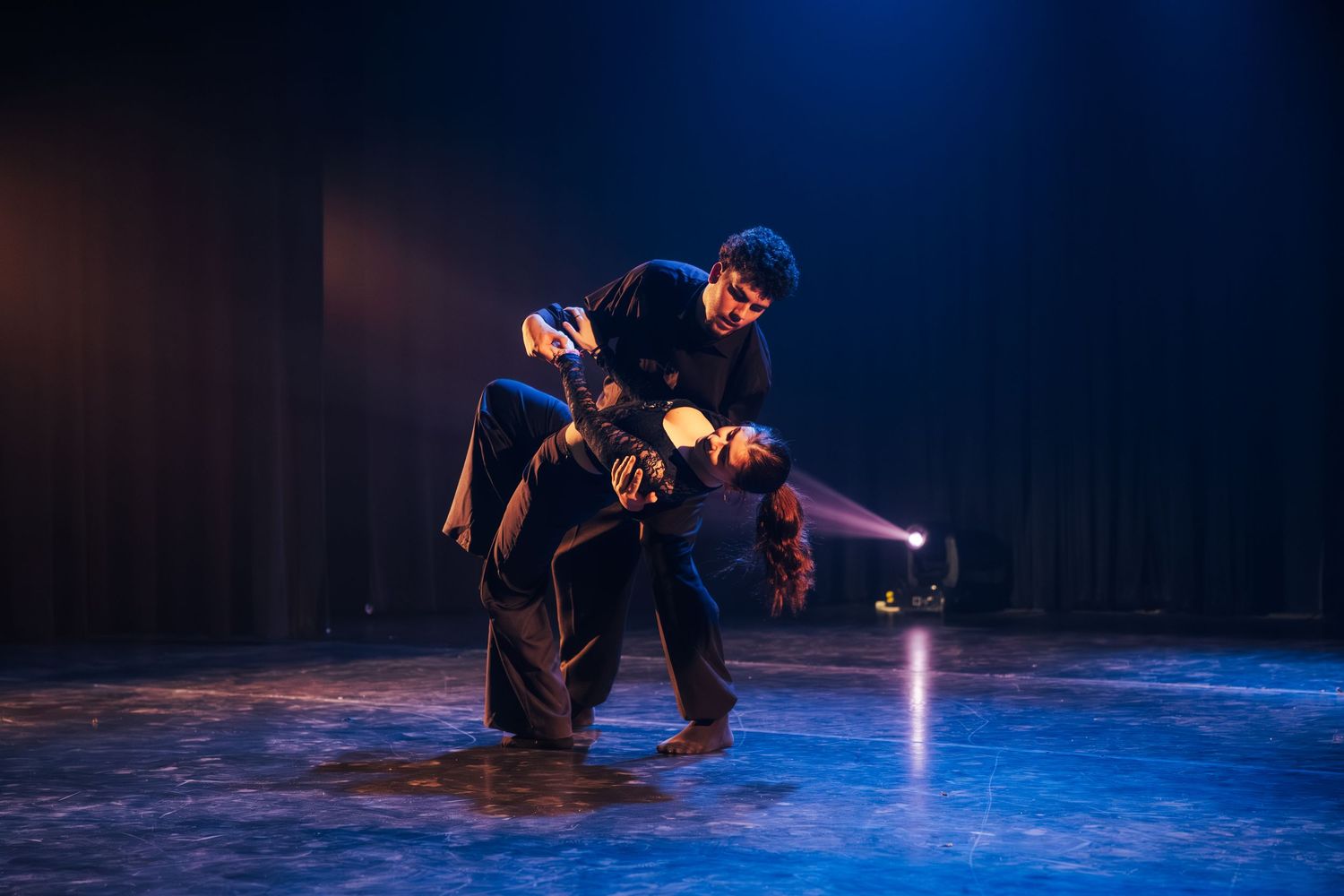 Performer in dark costume crouches dramatically on a dimly lit theater stage.