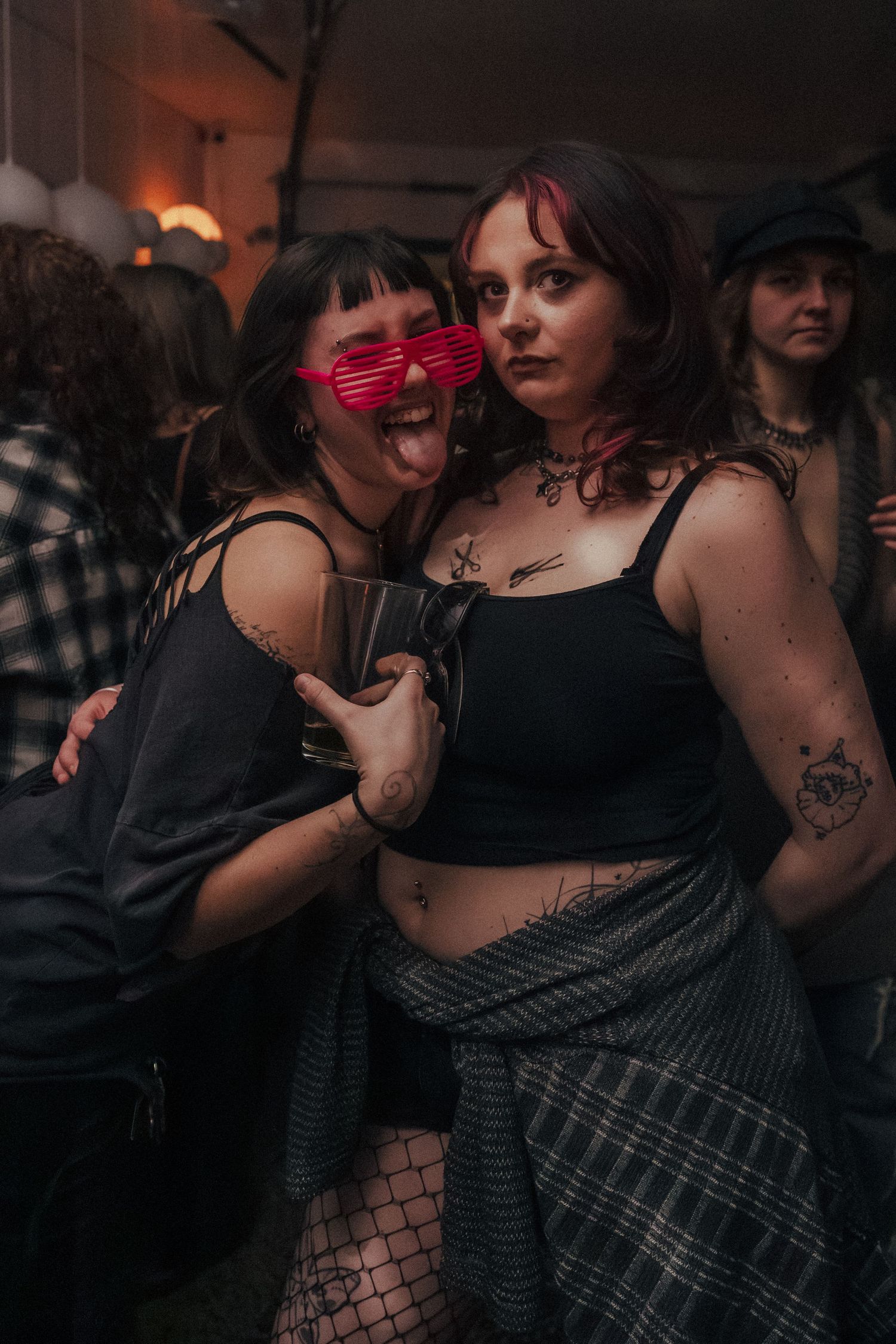 Two tattooed women pose together at a nightclub party, one wearing novelty pink shutter shades.