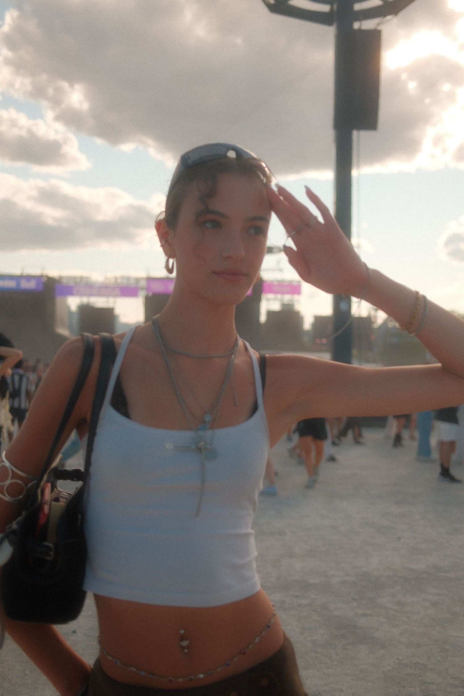 Young woman at outdoor music festival wearing white crop top, layered necklaces, and sunglasses on head.
