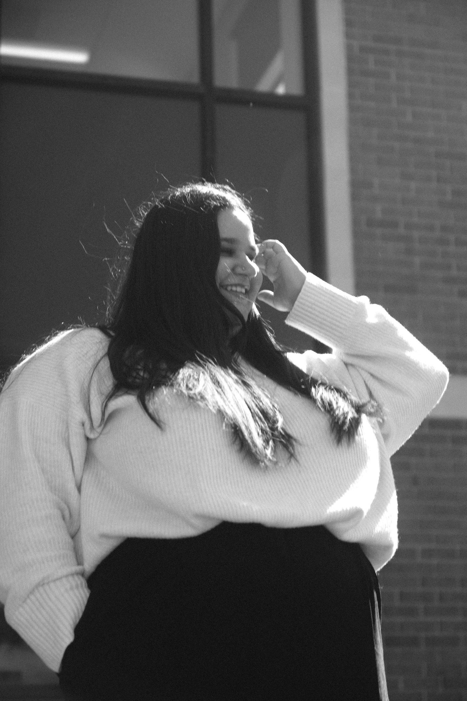Black and white portrait of a smiling young woman in a white sweater outdoors near a brick building.