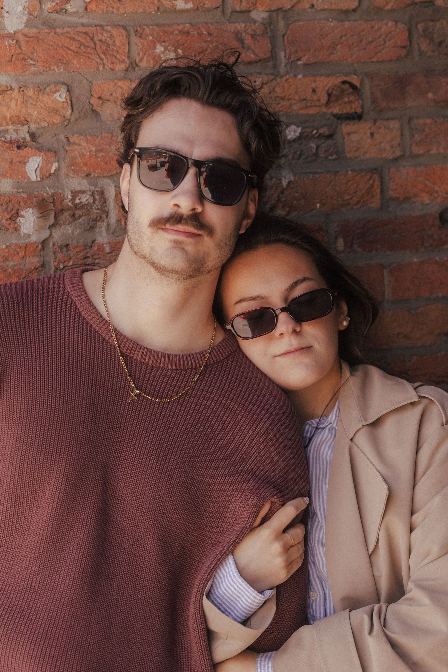 A stylish couple wearing sunglasses poses together against a rustic brick wall outdoors.