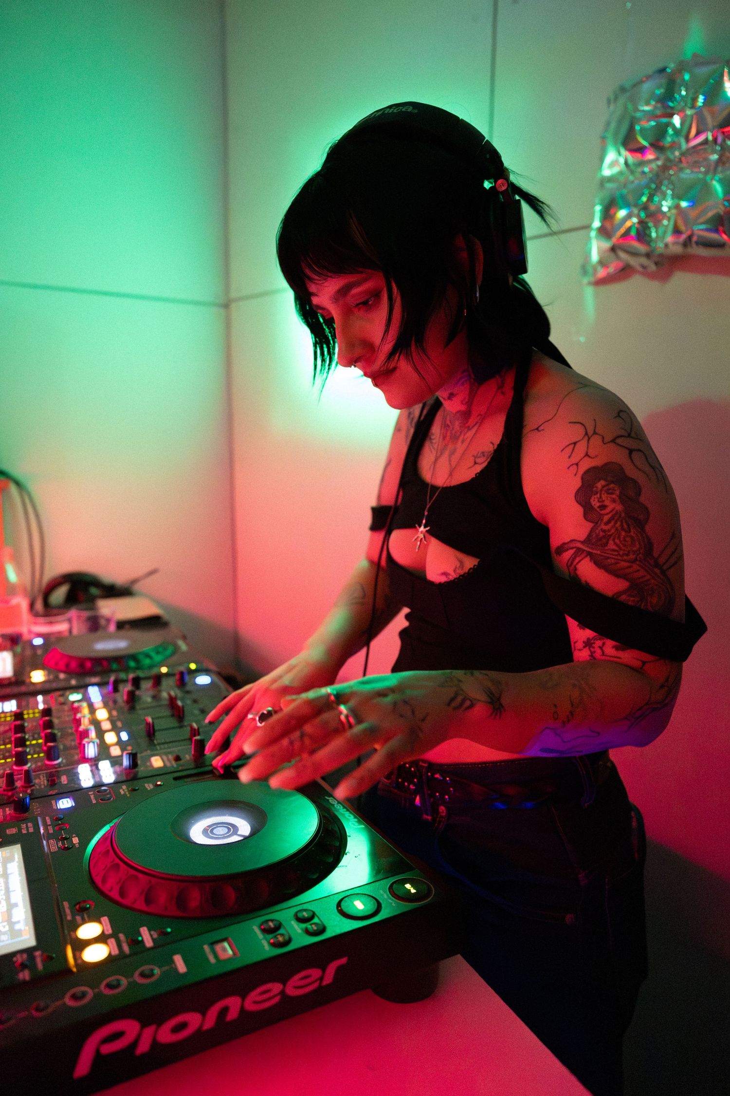 A female DJ with tattoos mixing music on Pioneer decks under green and pink lights.