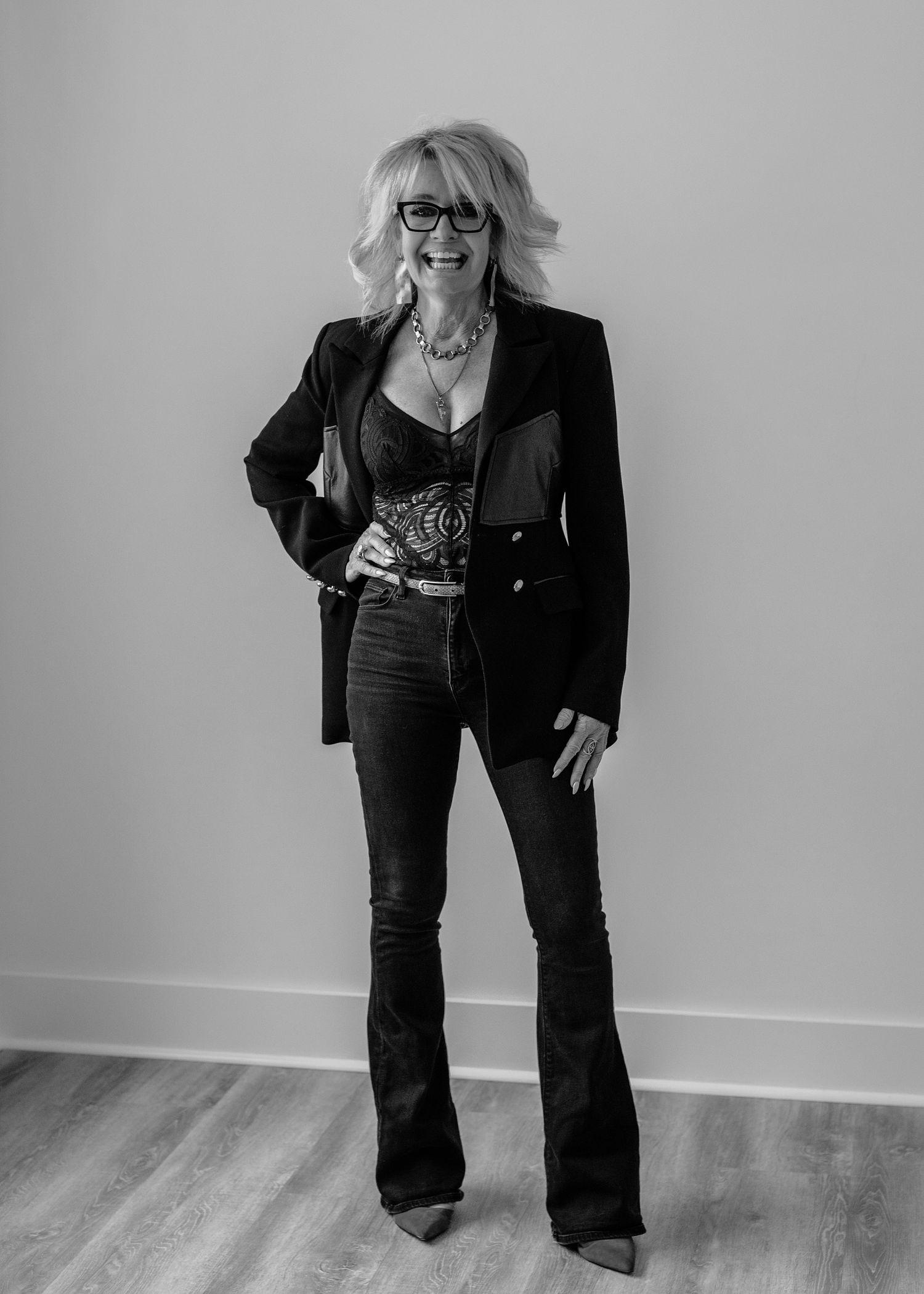 Stylish blonde woman in black blazer and flare jeans poses confidently, wearing glasses, black and white portrait.