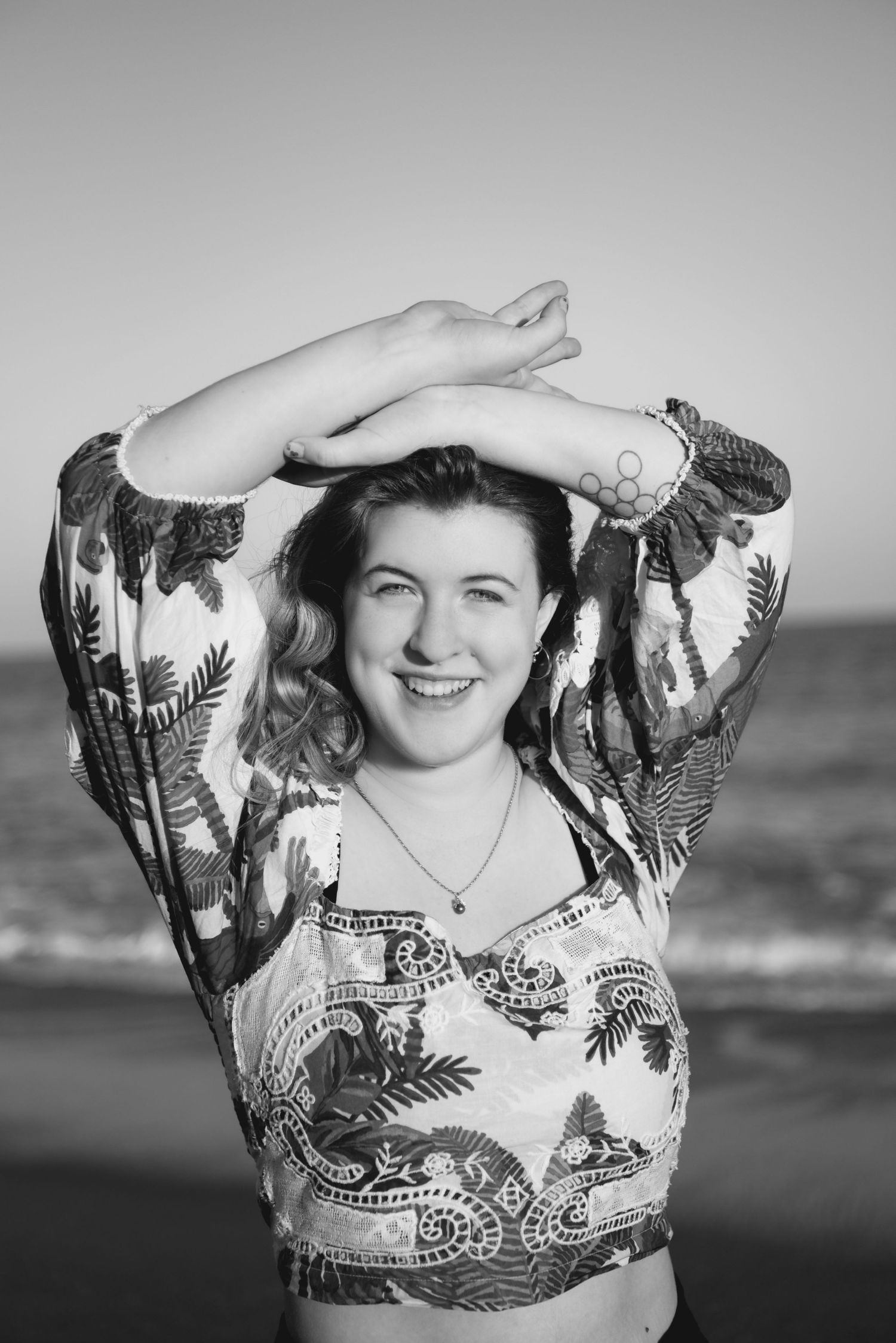 Black and white photo of a smiling woman with arms raised at the beach wearing a patterned crop top.