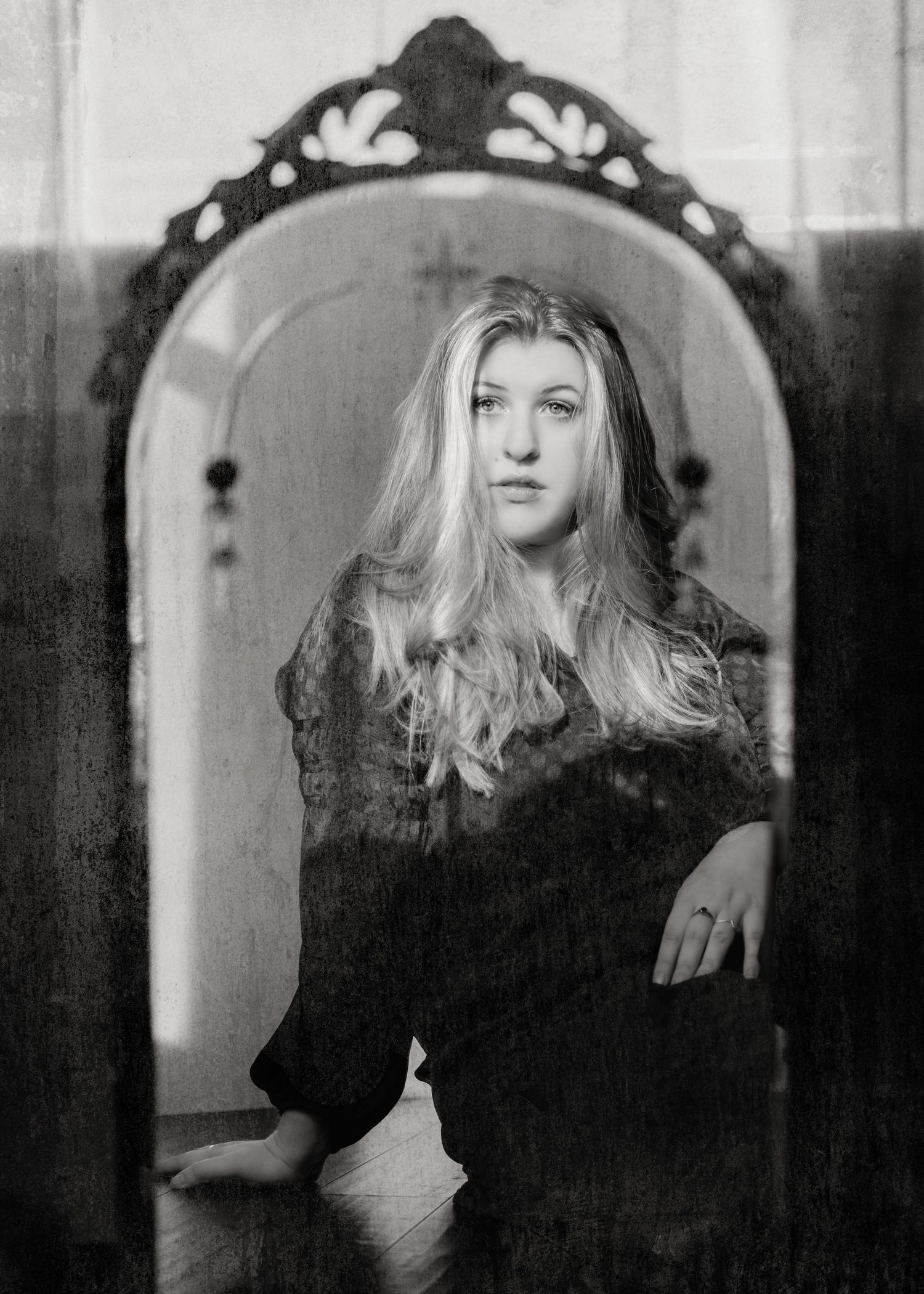 Black and white image of a blonde woman reflected in an ornate arched mirror seated on a floor.