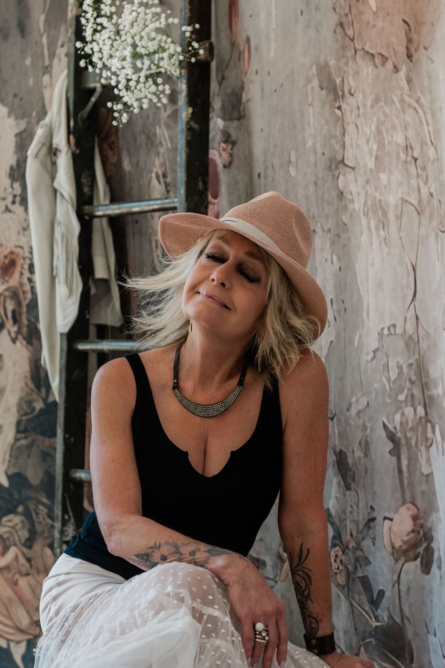 Blonde woman in black tank top and wide-brim beige hat with silver necklace sits against rustic floral wall.