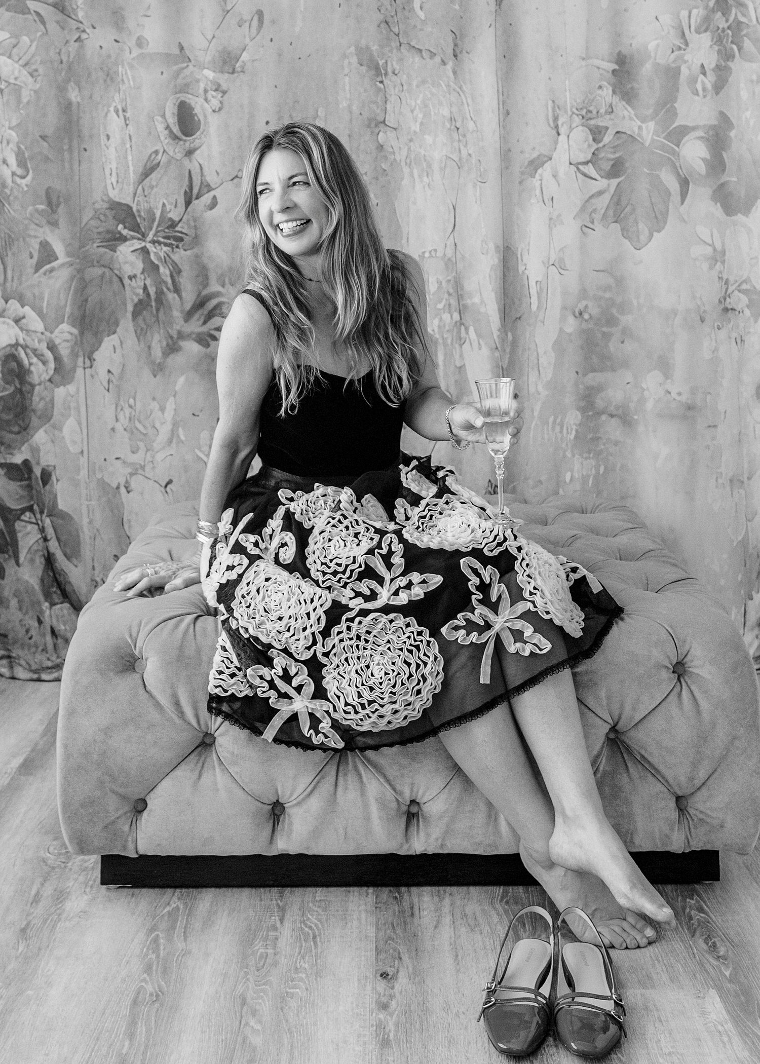 Woman in floral skirt sits on tufted sofa holding wine glass, smiling, shoes on floor, black and white photo.