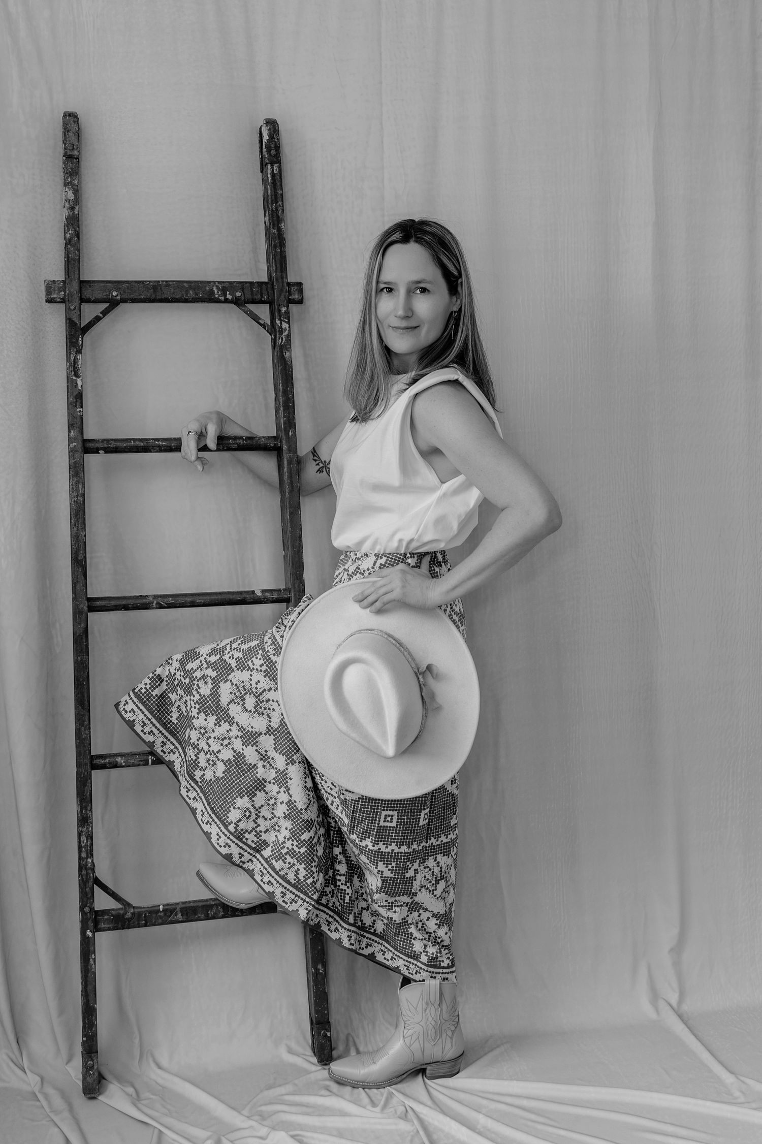 Black and white photo of woman in white top and patterned skirt holding cowboy hat beside rustic wooden ladder.