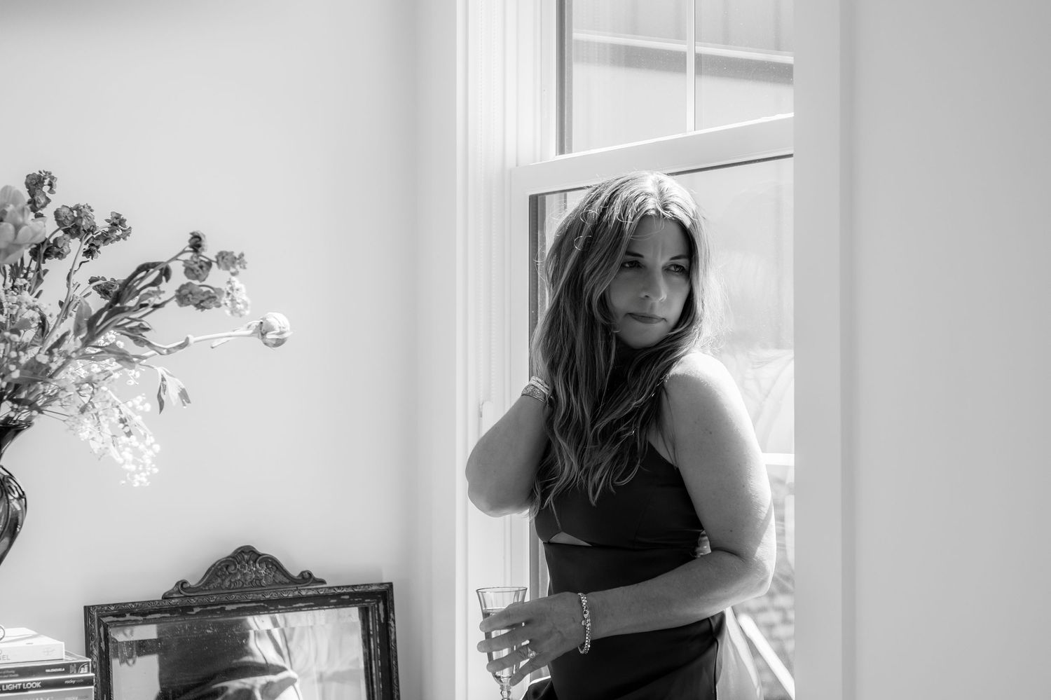 Black and white photo of brunette woman in black dress standing by sunlit window with flowers nearby.