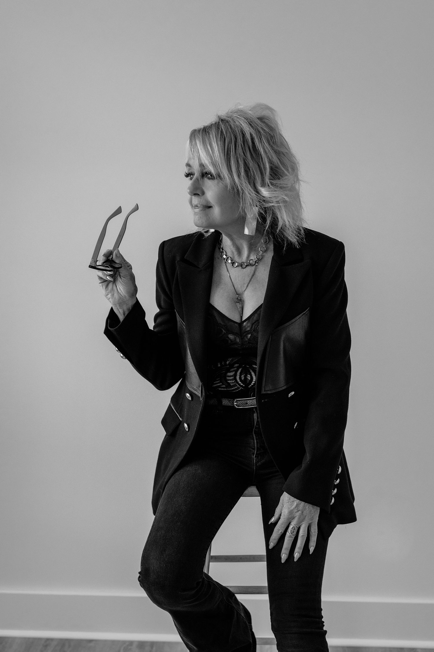 Black and white photo of blonde woman in black blazer and jeans holding eyeglasses while sitting on stool.