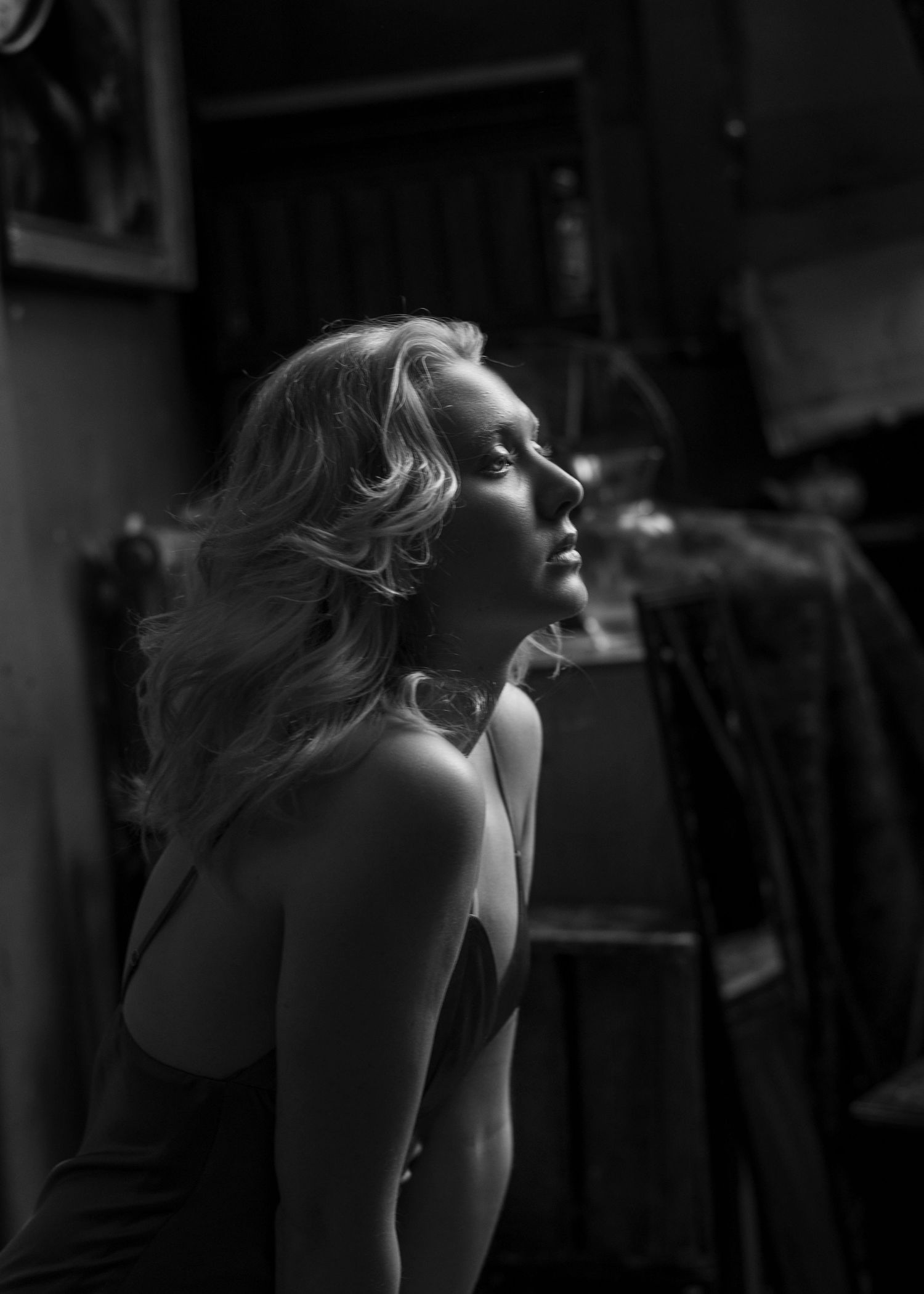 Dramatic black and white side profile of a blonde woman in a sleeveless dress gazing upward in a dark room.