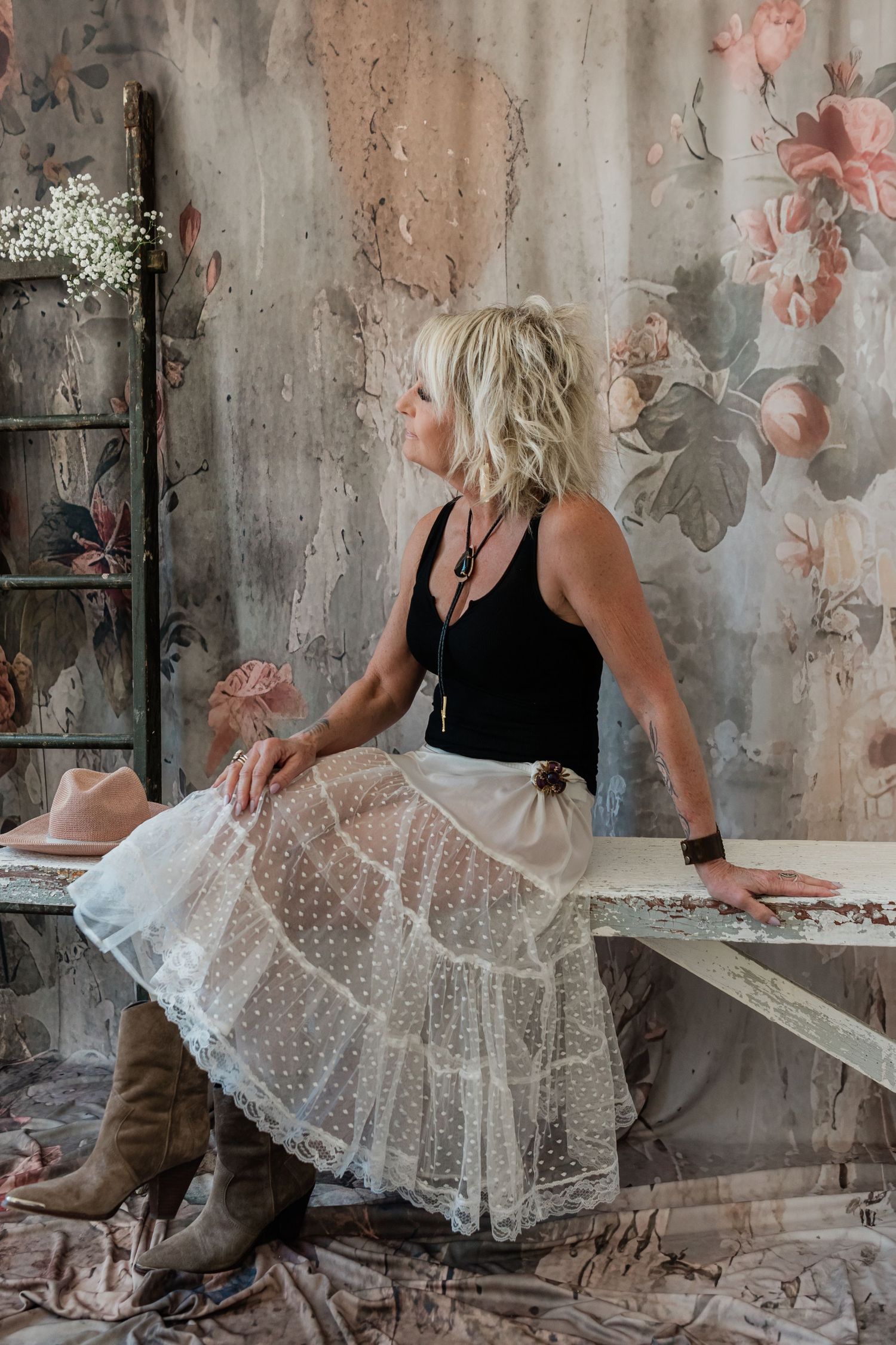 Blonde woman in lace skirt and black top sits on bench before floral wallpaper, gazing away, color photo.