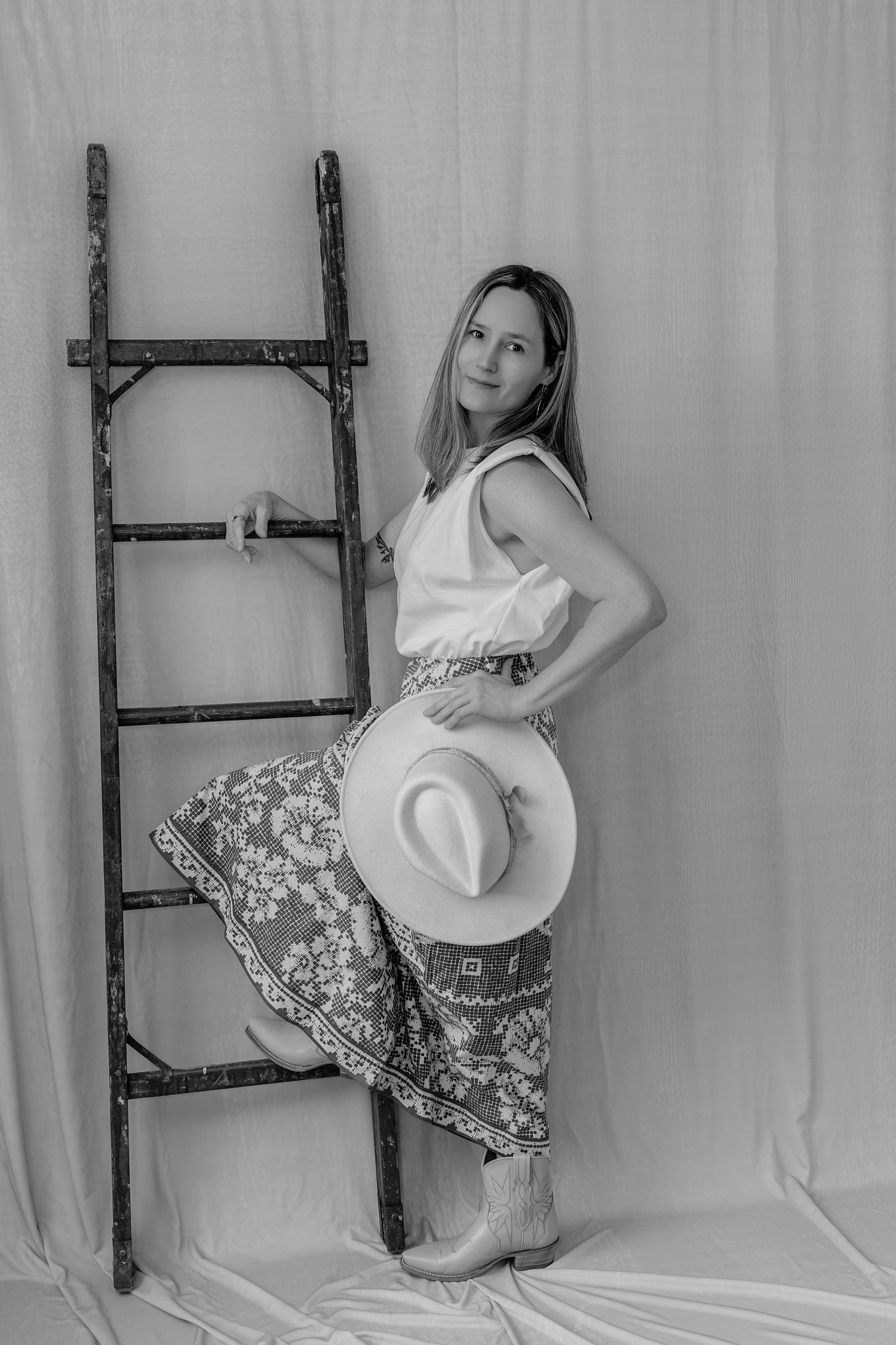 Woman in white top and floral pants poses beside rustic ladder holding cowboy hat, black and white studio shot.