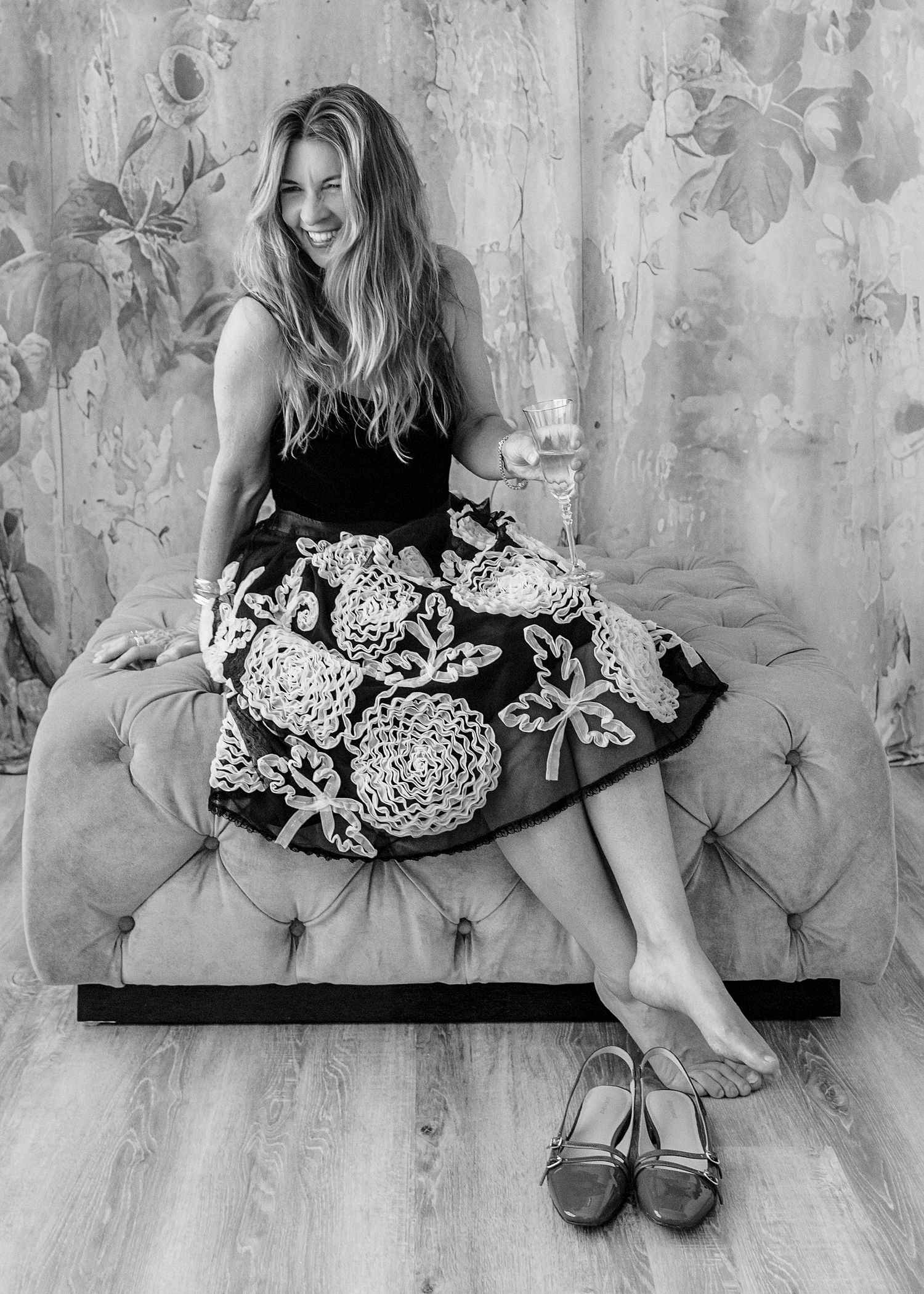 Laughing woman in embroidered skirt seated on tufted couch with wine glass, heels on floor, black and white.