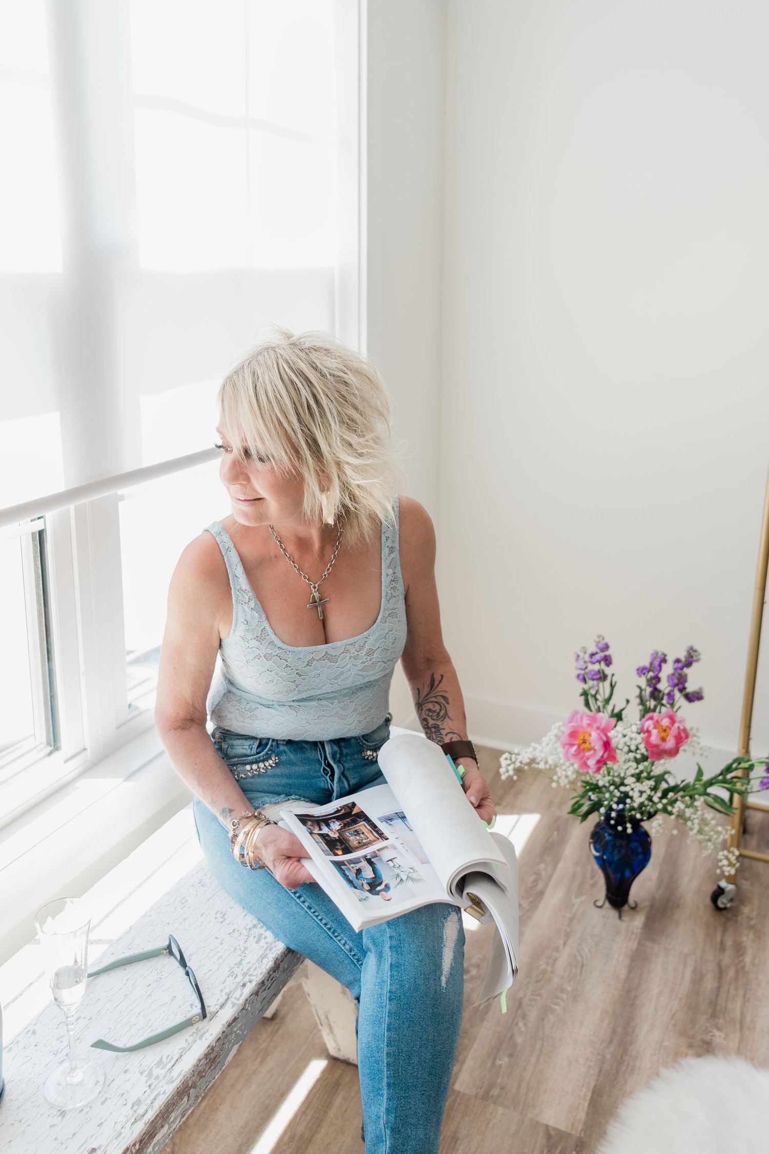 Blonde woman in blue tank top and jeans reads magazine while sitting by bright window with flowers nearby.