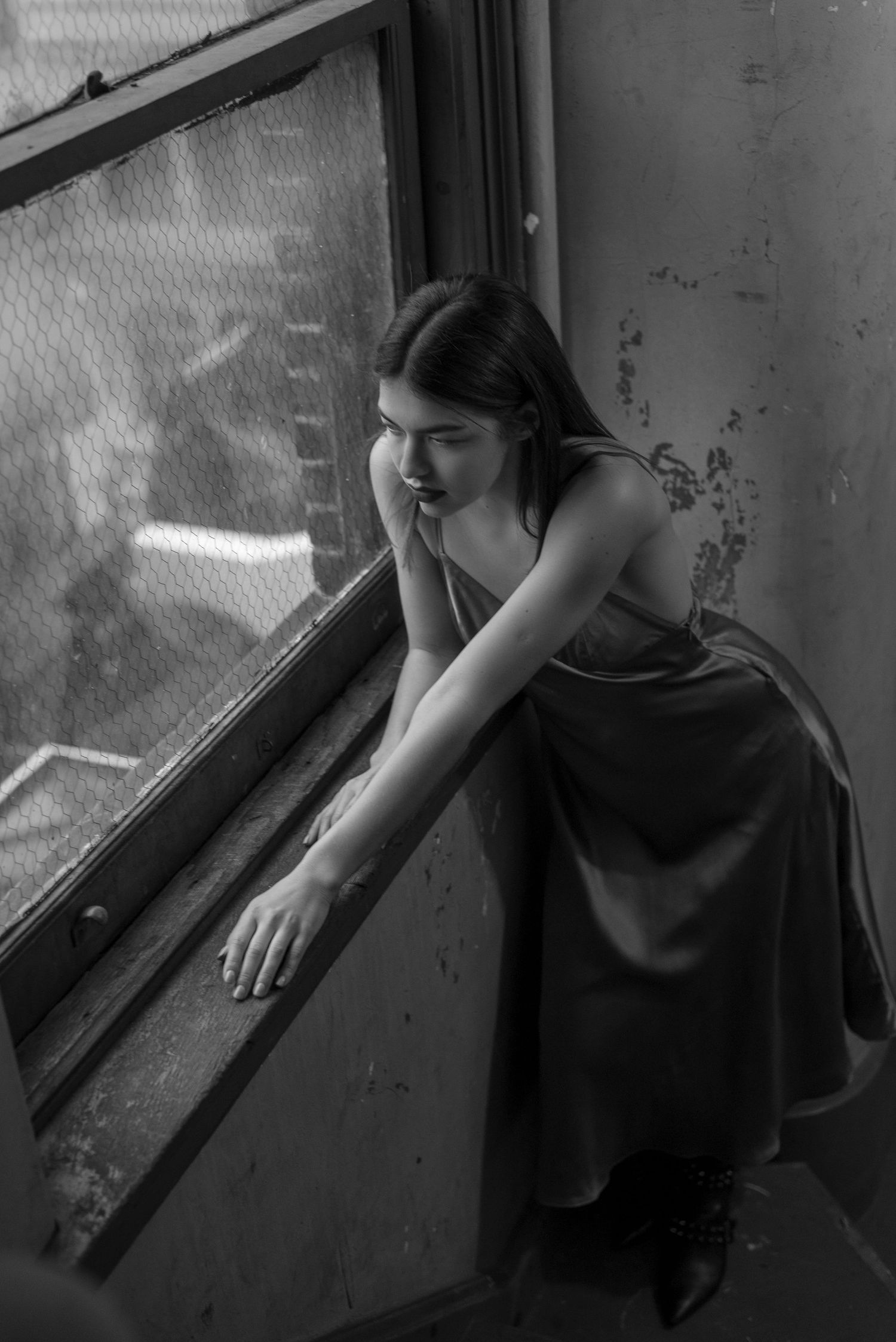 Dark-haired woman in satin dress leaning on open window in black and white urban setting.