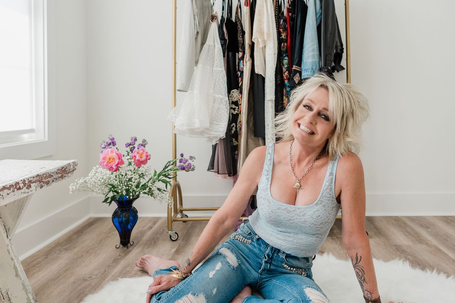 Smiling blonde woman in blue tank and ripped jeans sits on white rug near clothing rack and flowers.