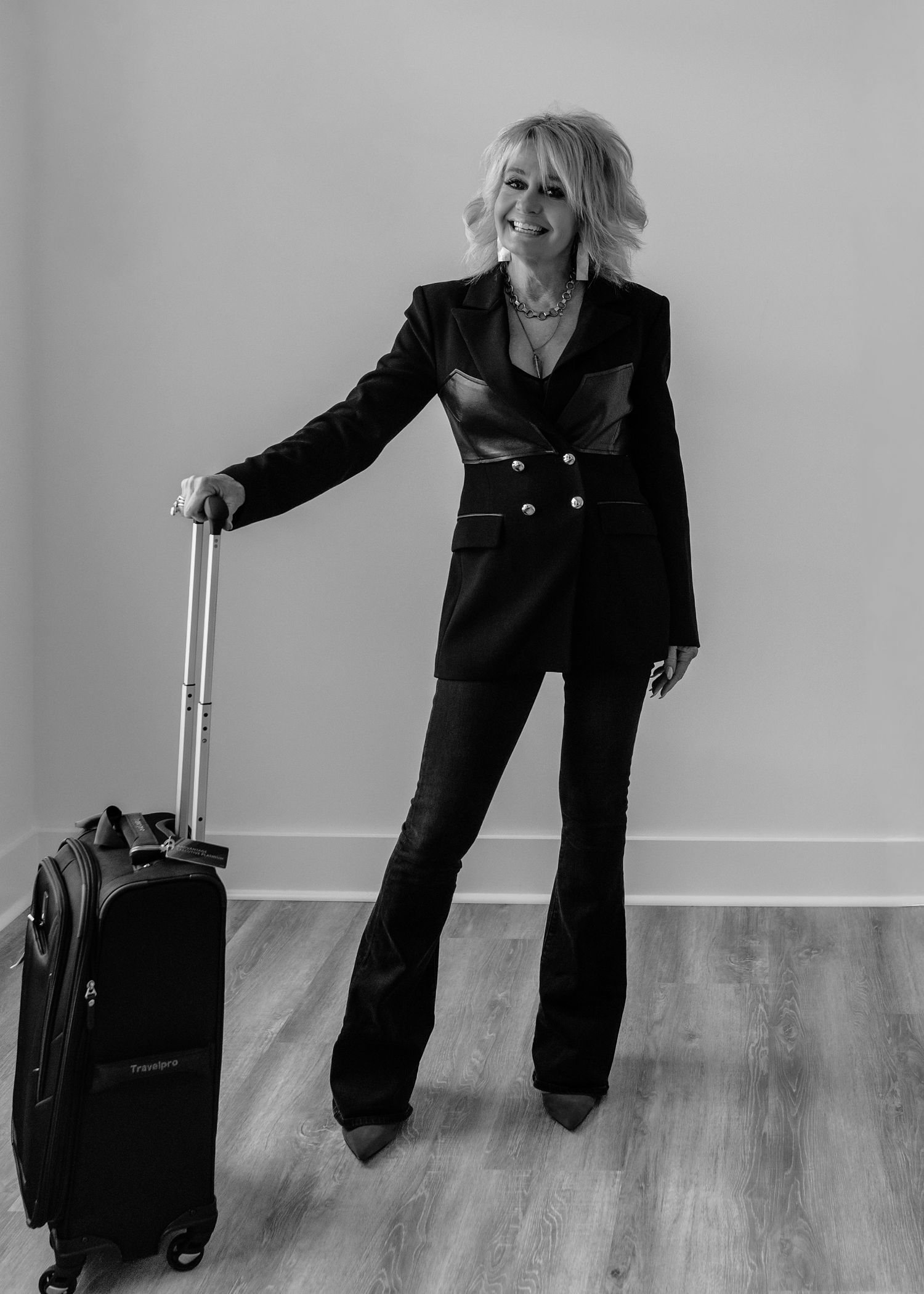 Black and white photo of smiling blonde woman in black blazer and flare pants posing with rolling luggage.