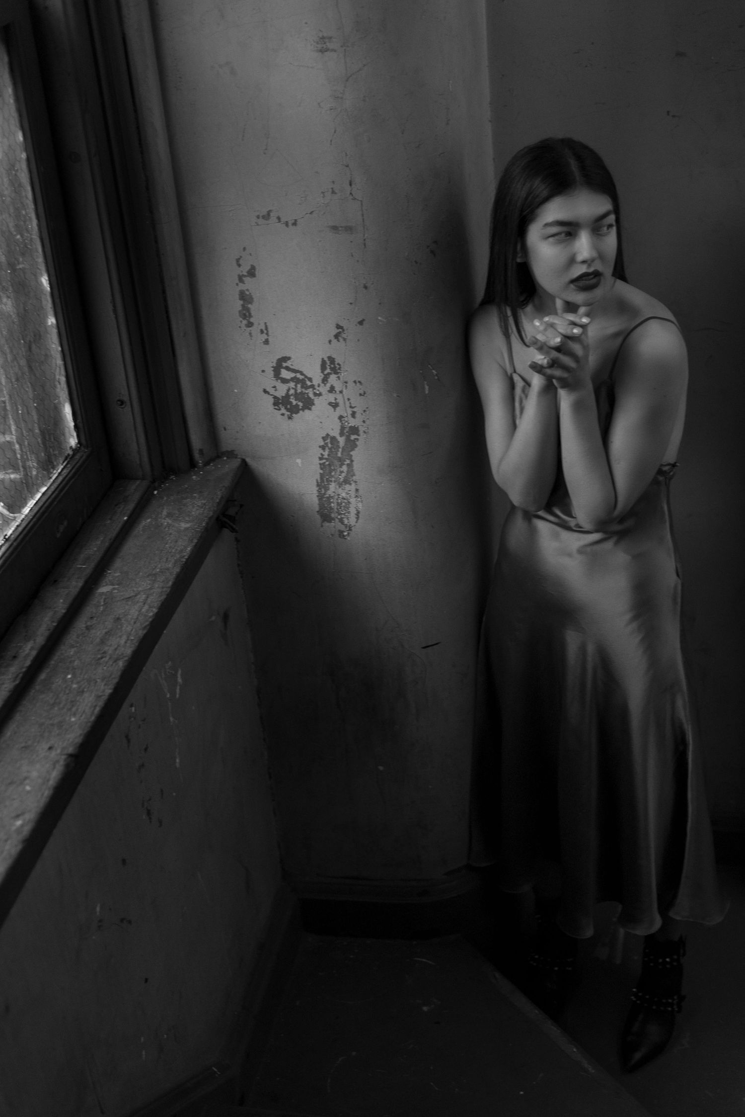 Young woman in satin slip dress standing by window in moody black and white portrait.