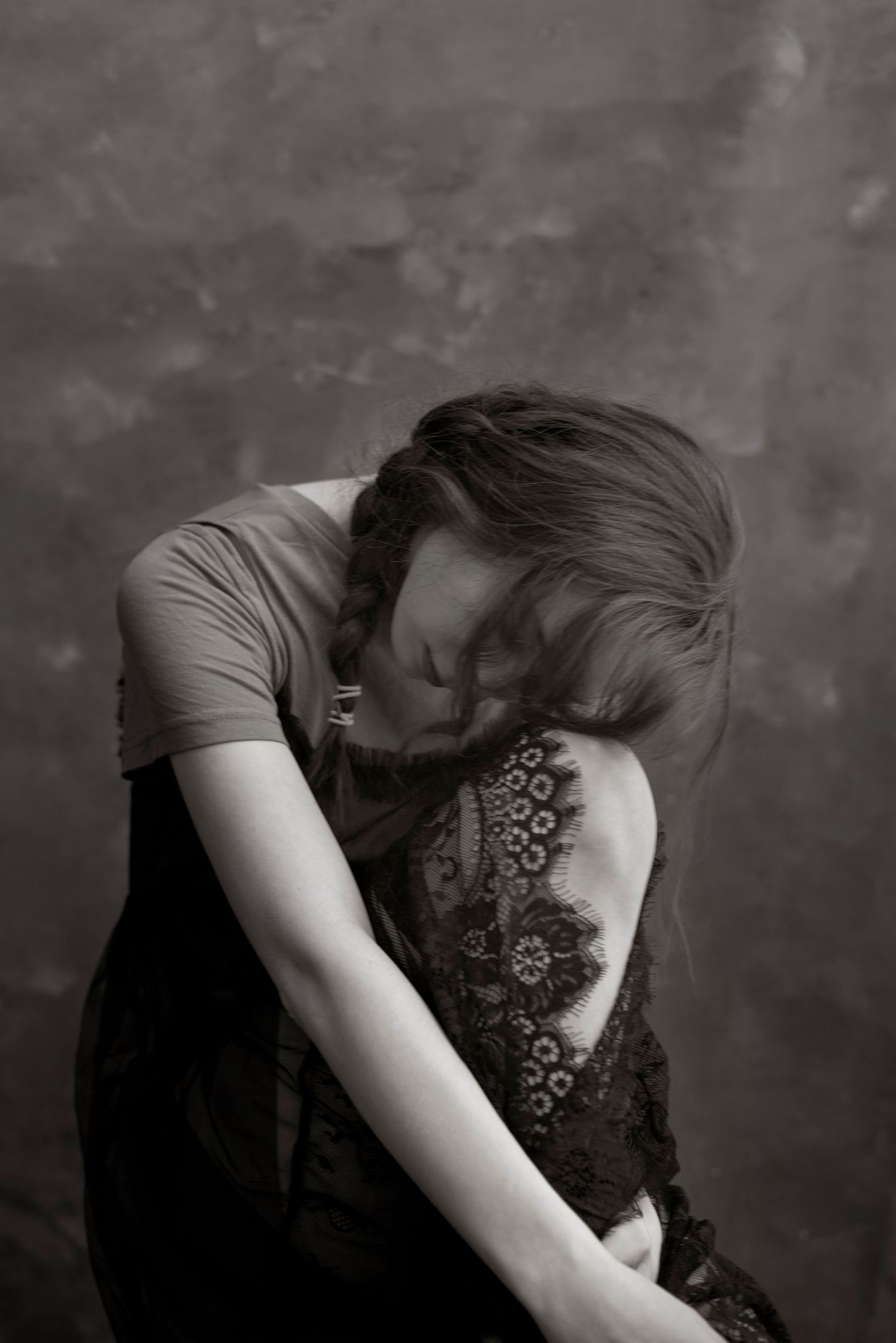 Black and white photo of a woman with braided hair hugging her knees, head bowed, against a gray wall.
