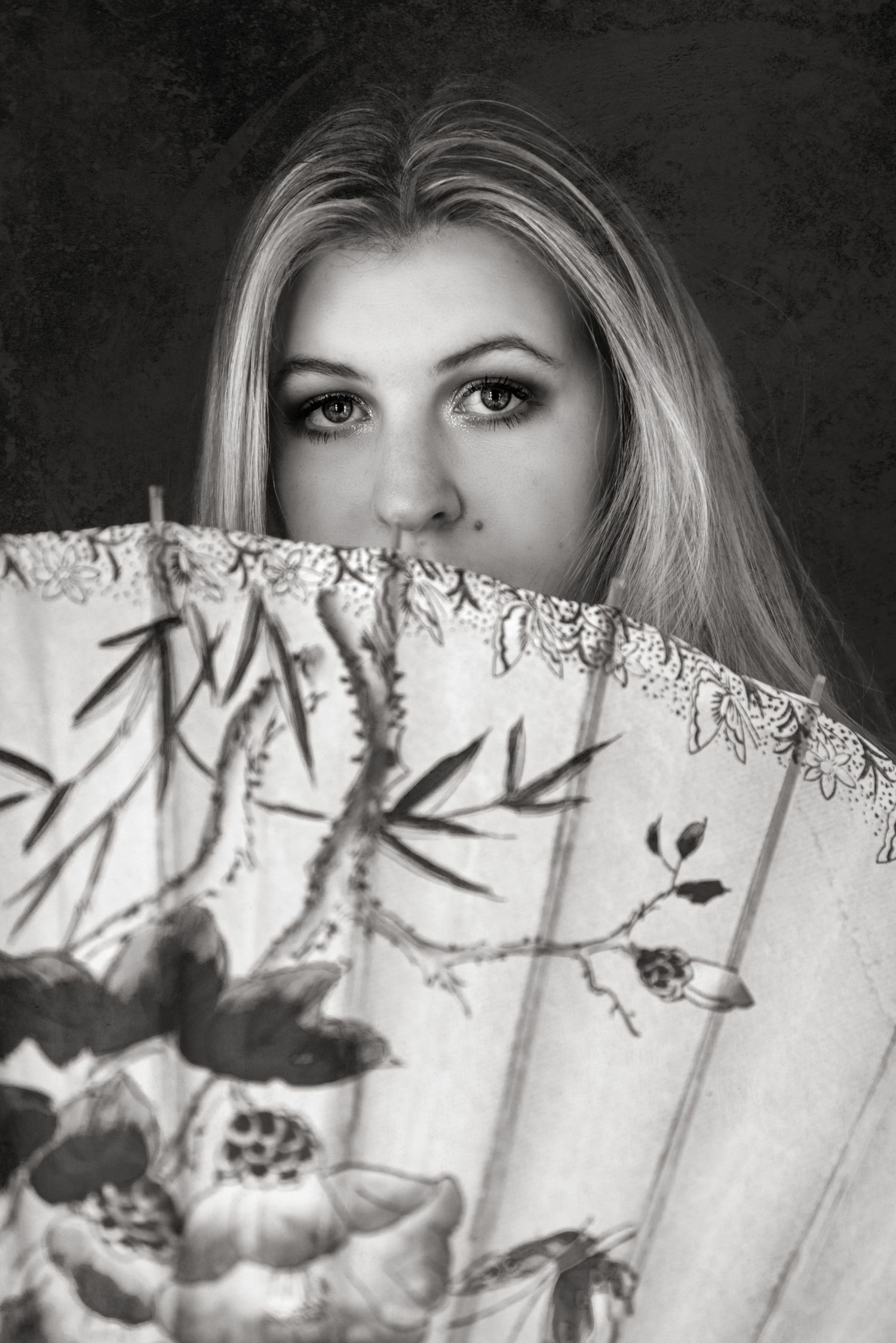 Blonde woman partially hiding her face behind a decorative floral hand fan in a black and white portrait.