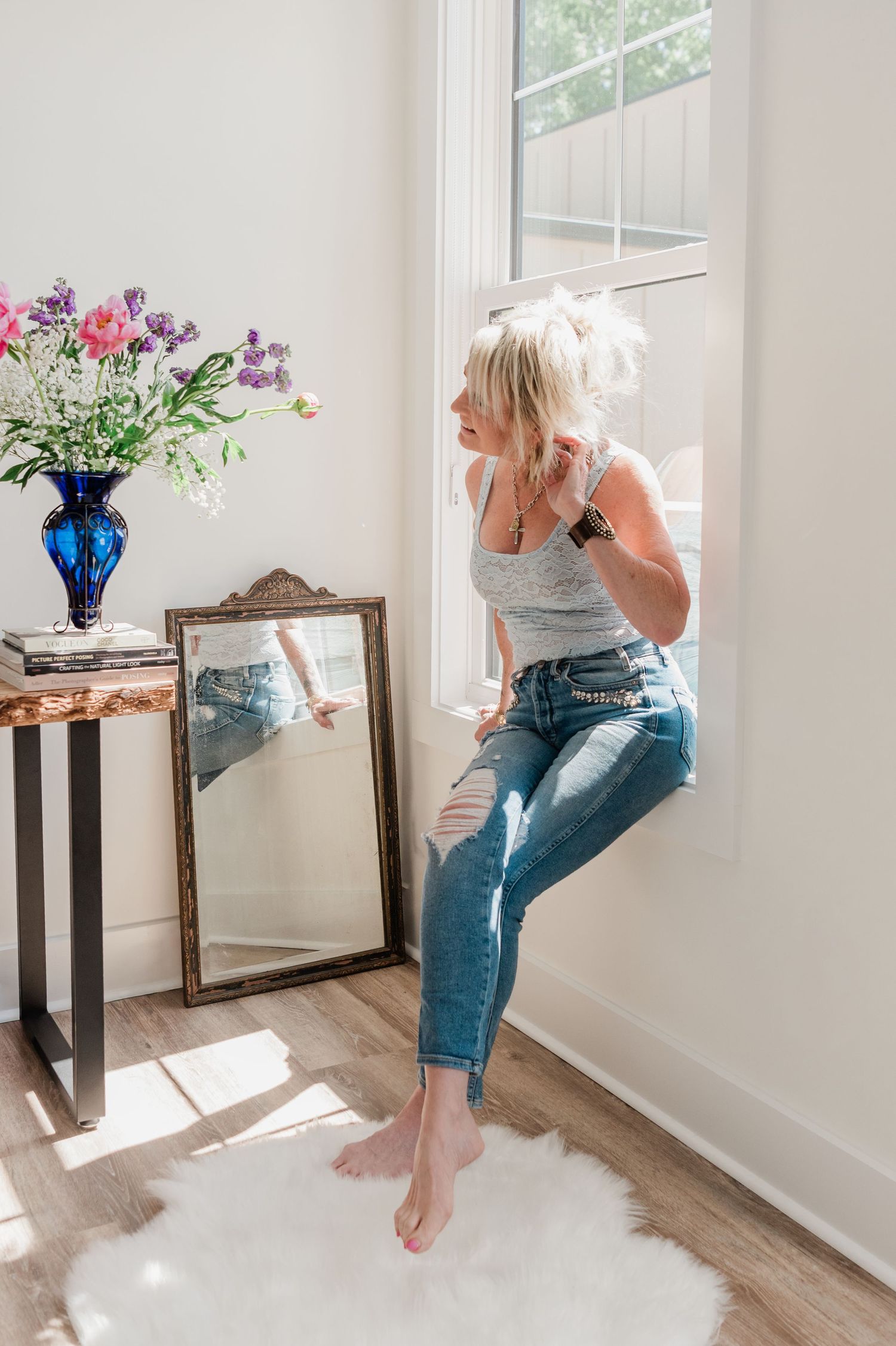 Blonde woman in gray tank and ripped jeans sits on window ledge beside flowers and mirror, color photo.