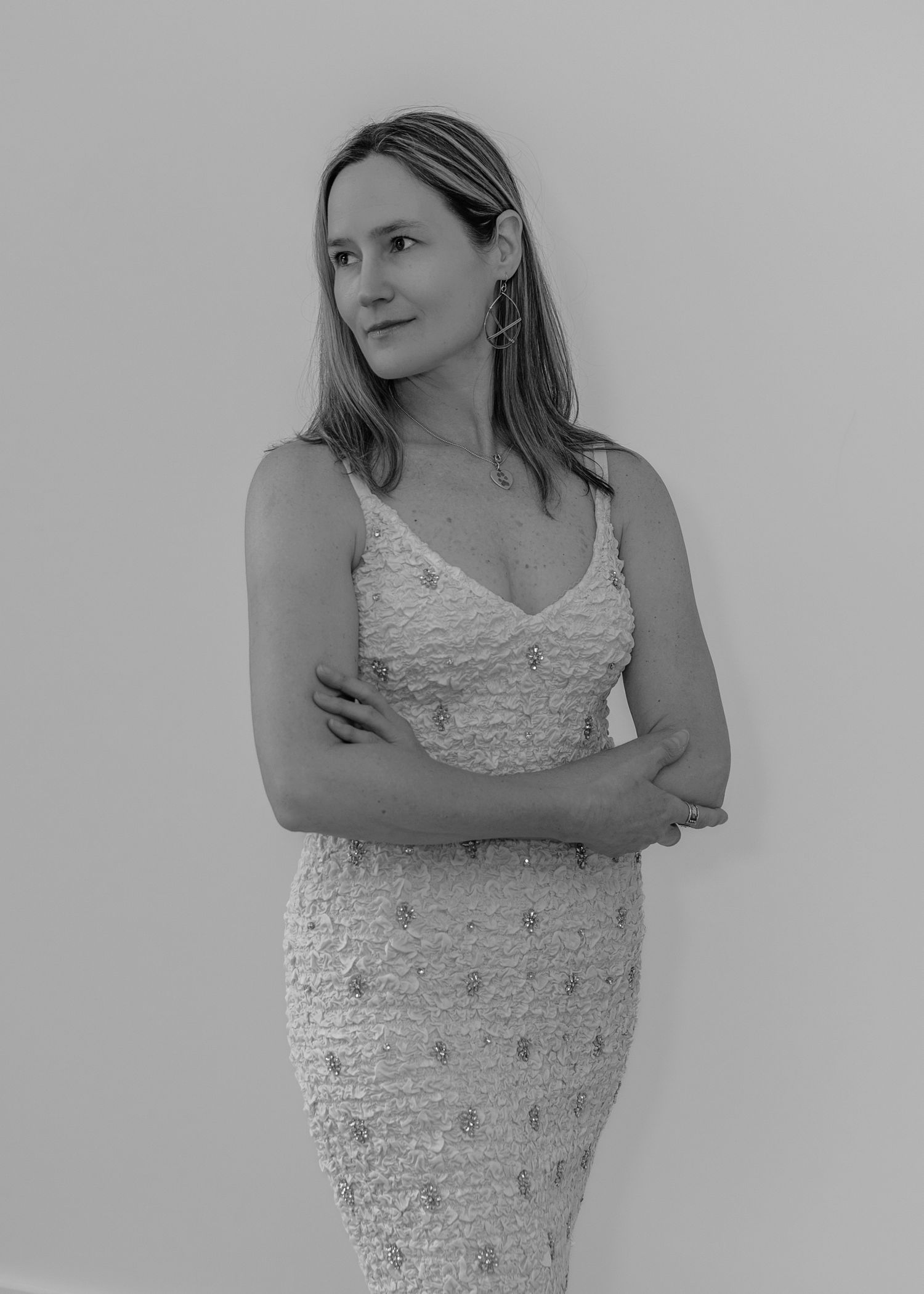 Elegant woman in beaded spaghetti strap dress stands with arms crossed, looking away, black and white photo.