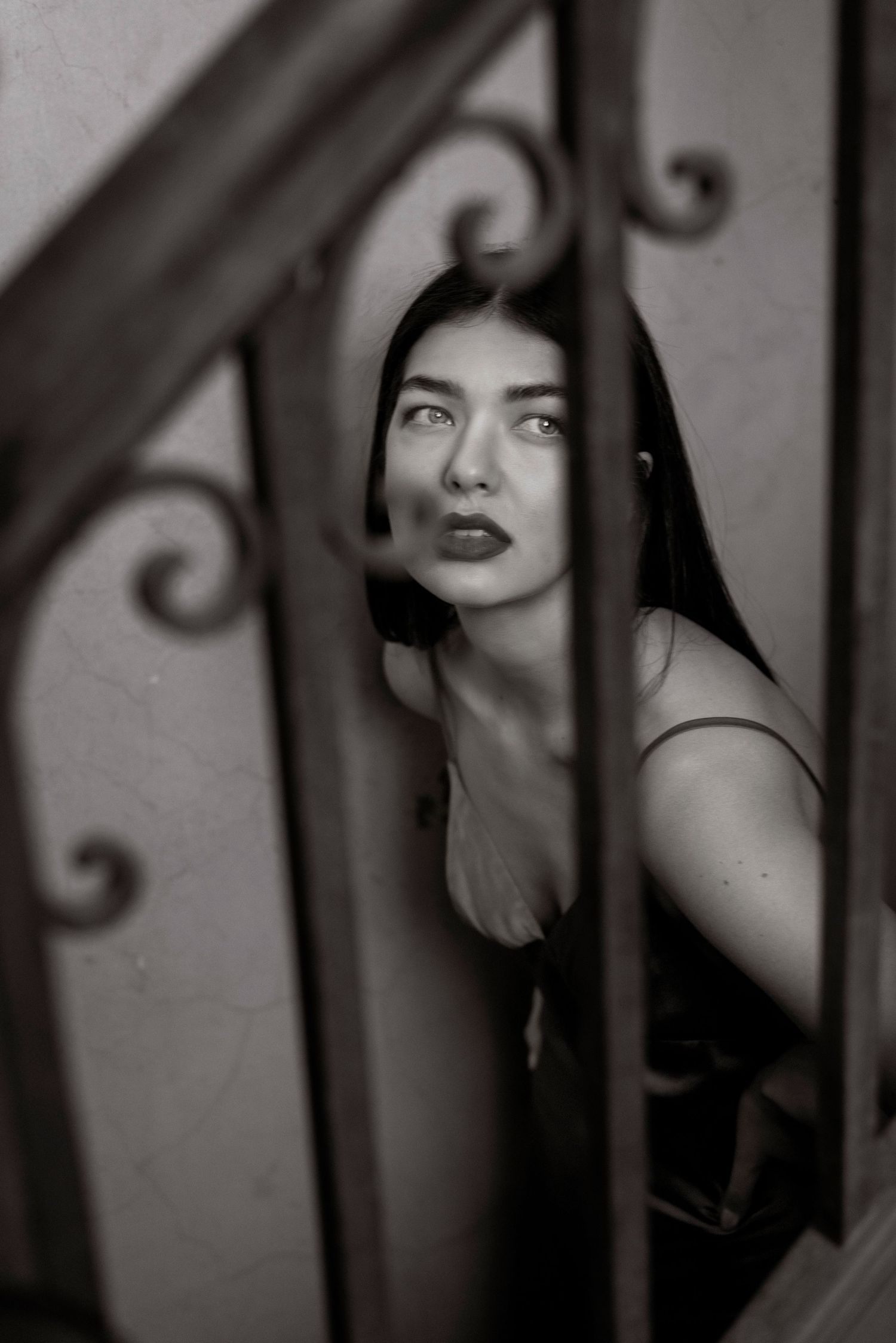 Dramatic black and white photo of a dark-haired woman peering through ornate wrought iron stair railings.
