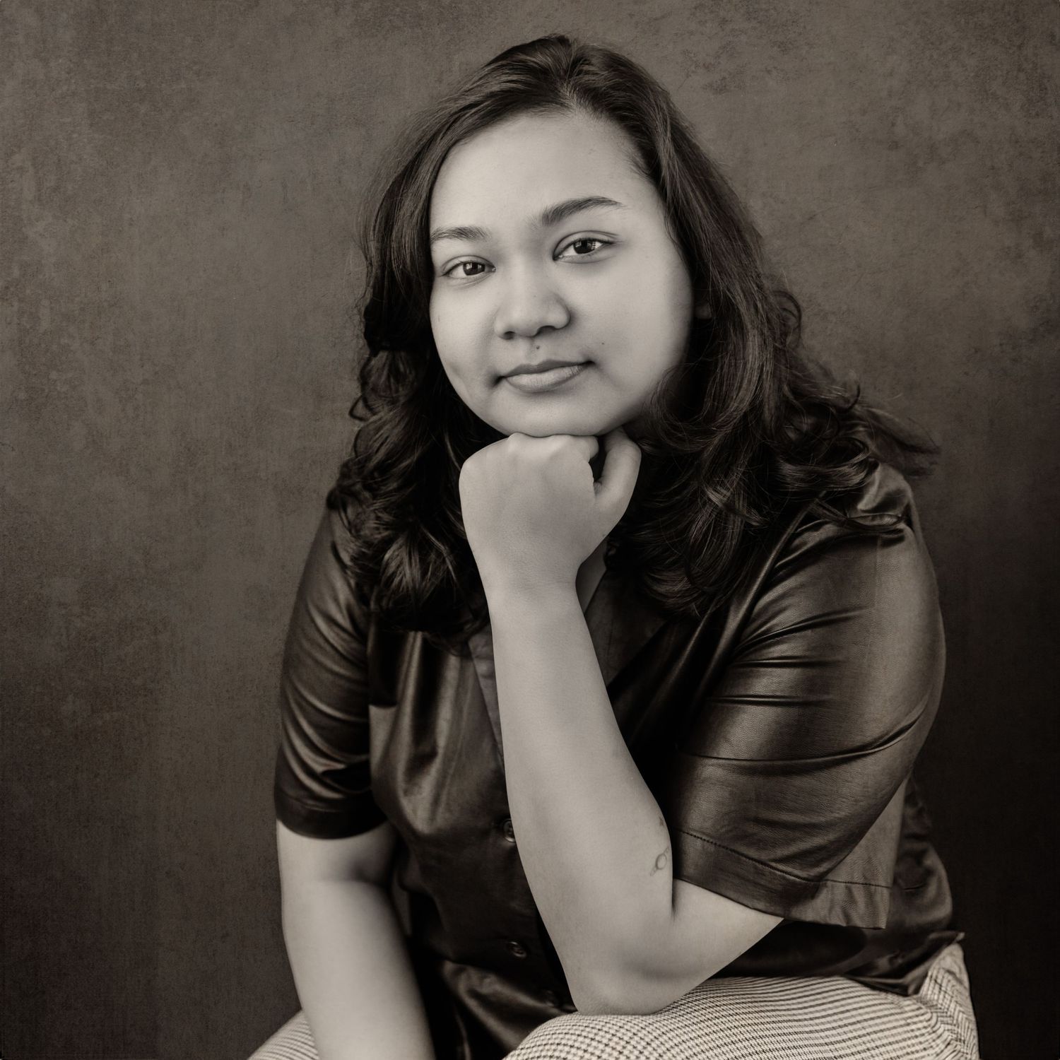 Young woman with dark hair resting chin on hand, smiling in a black and white studio portrait.