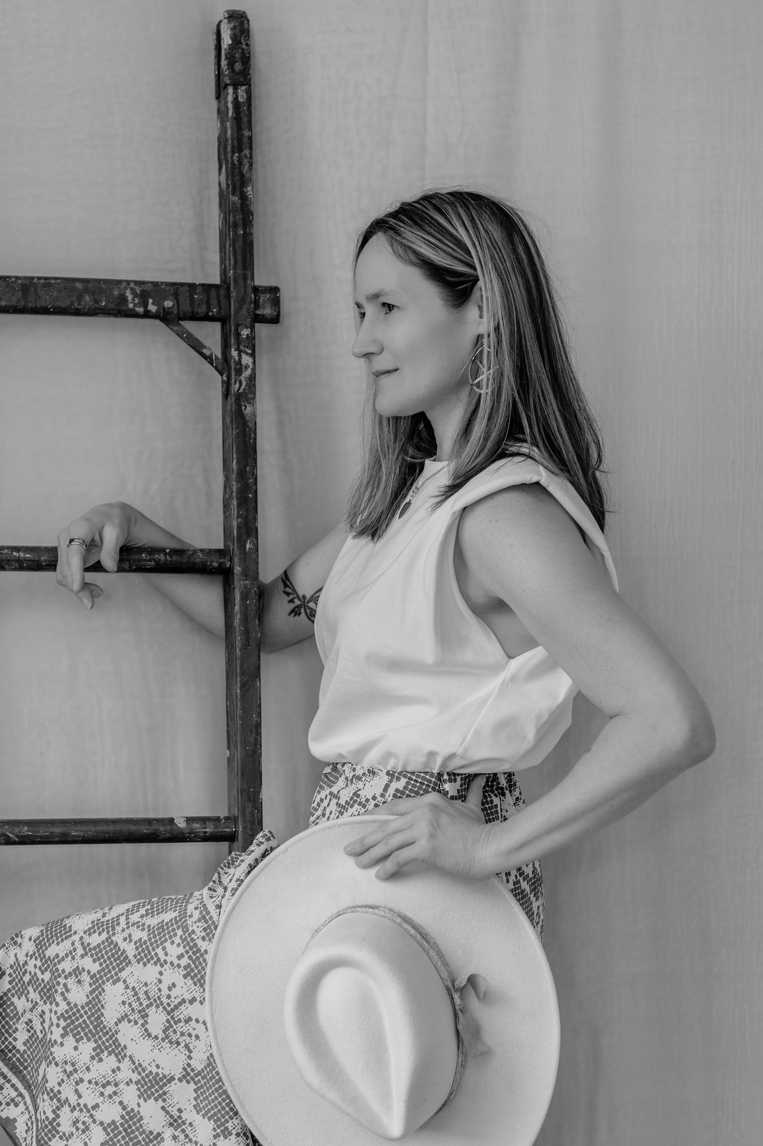 Black and white portrait of a woman in a white top holding a hat beside a rustic wooden ladder.