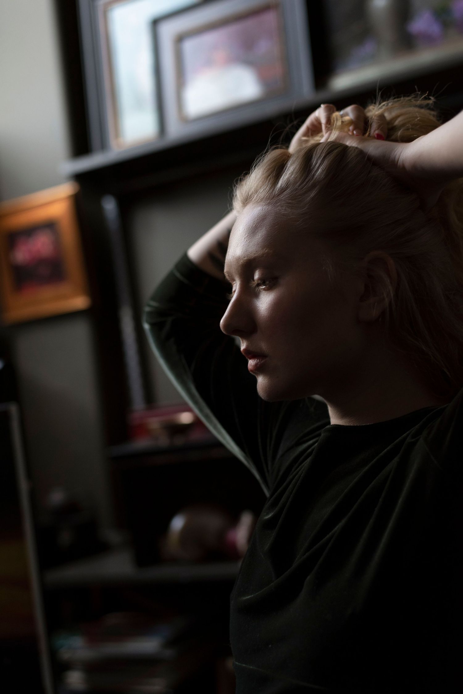 Side profile of a blonde woman pulling her hair back in a dimly lit room with shelves behind her.