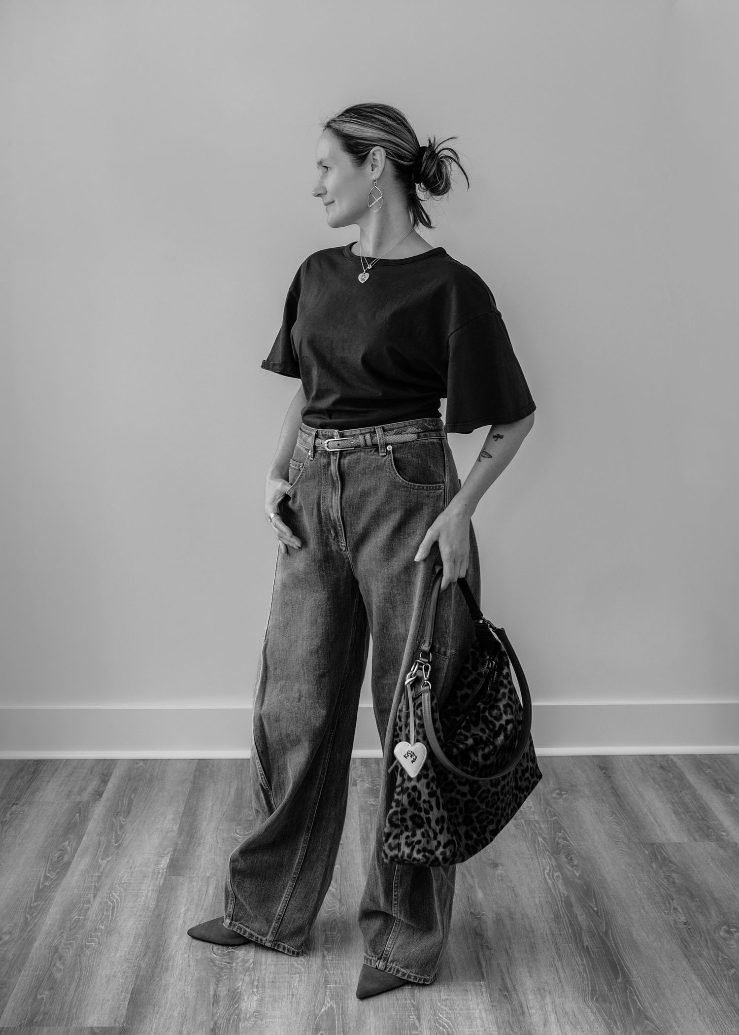 Stylish woman in black tee and wide-leg jeans holds leopard print bag, looking sideways, black and white photo.