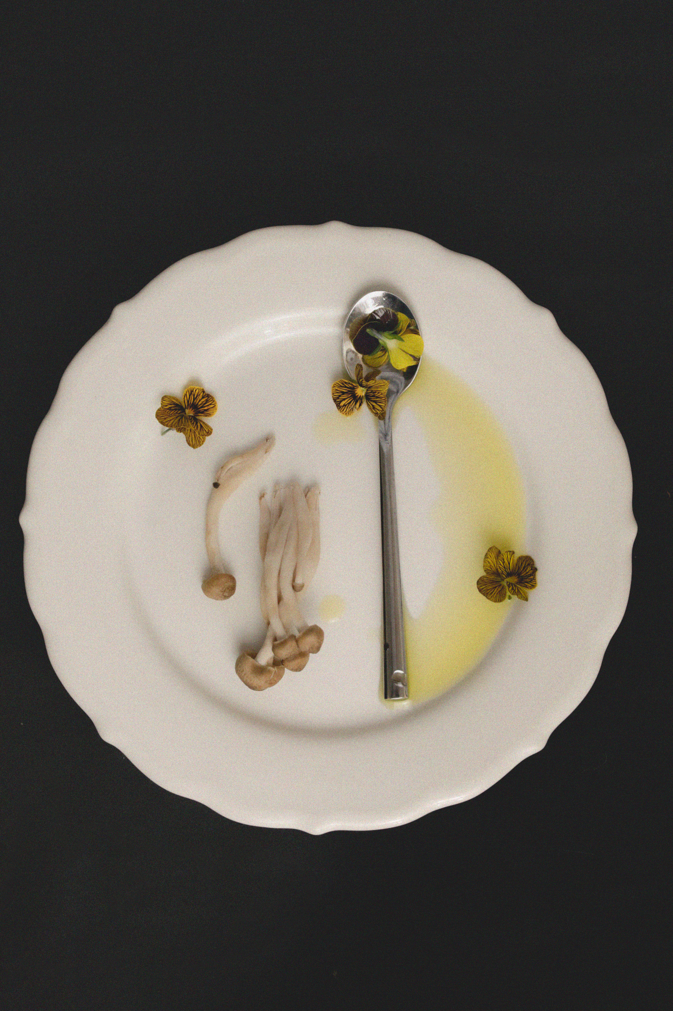 White ceramic plate with enoki mushrooms, yellow viola flowers, and a silver spoon with olive oil on black background.