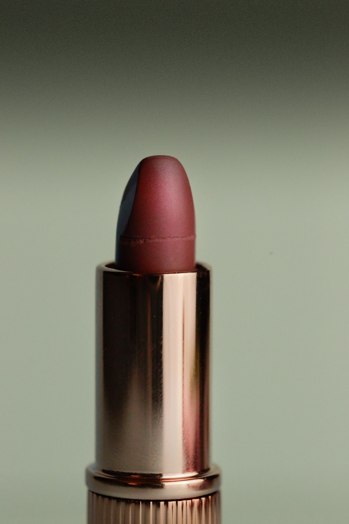 Close-up of an open rose-gold tube of matte mauve-pink lipstick against a soft green background.