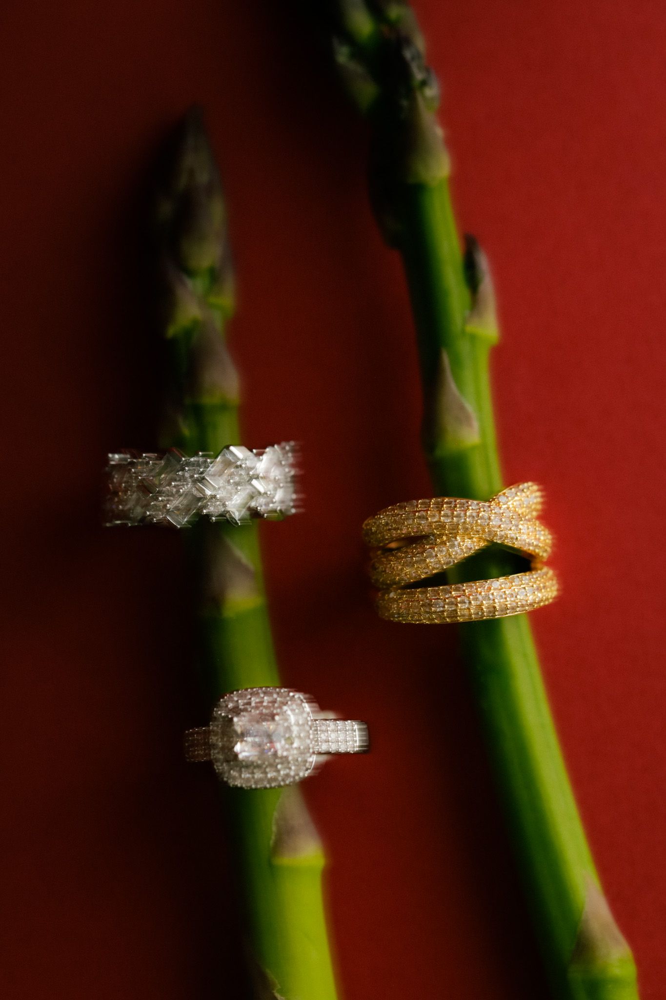 Three diamond rings displayed on green asparagus stalks against a rich red background, featuring white gold and yellow gold designs.