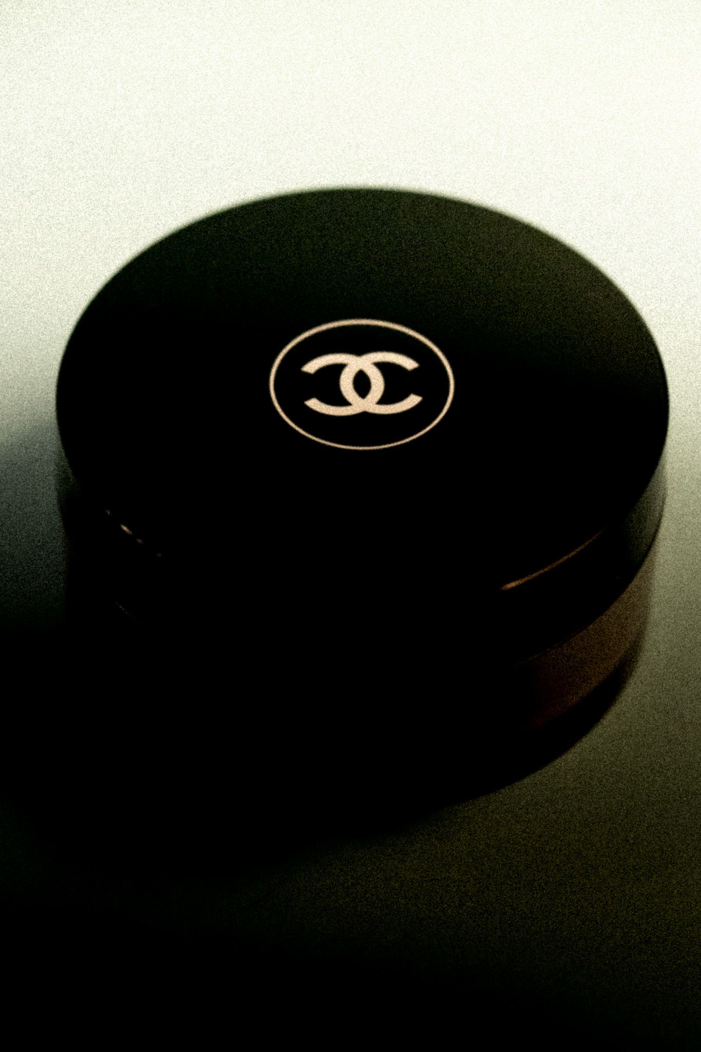 Chanel black round compact powder case with iconic interlocking CC logo on dark reflective surface.