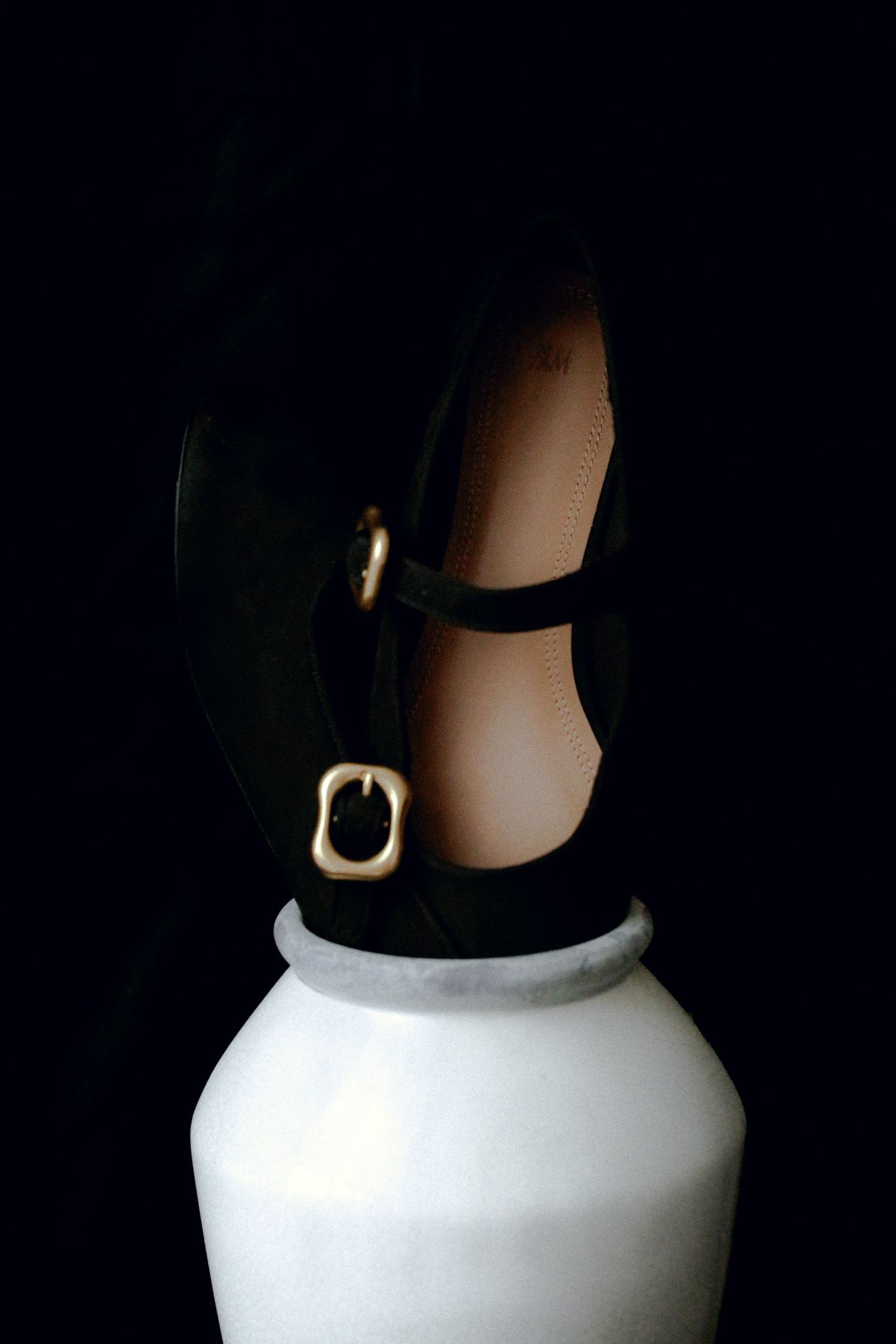 Black suede Mary Jane shoe with gold buckle strap balanced on white ceramic vase against dark background.