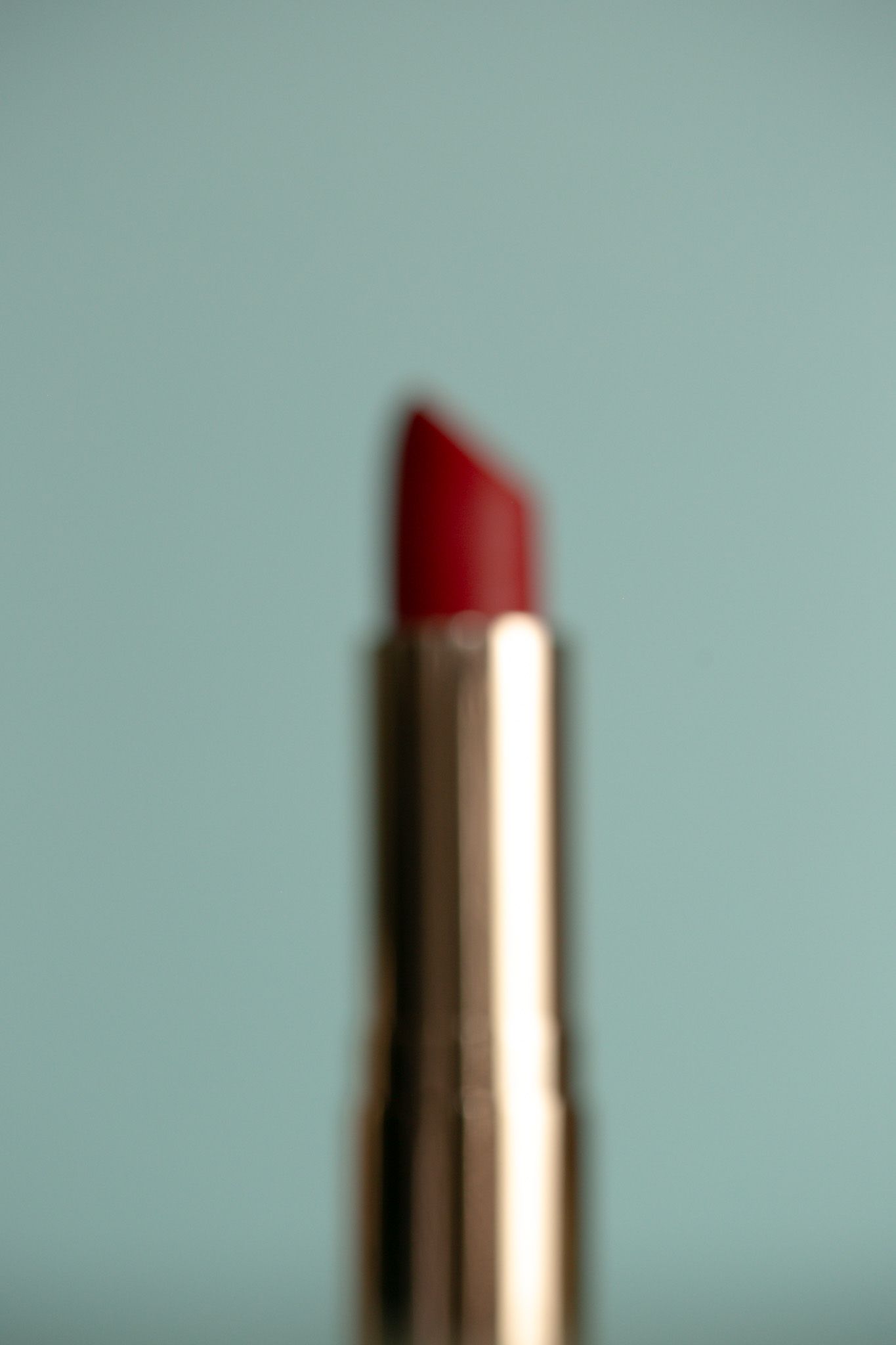A blurred red lipstick in a gold tube against a soft teal background.