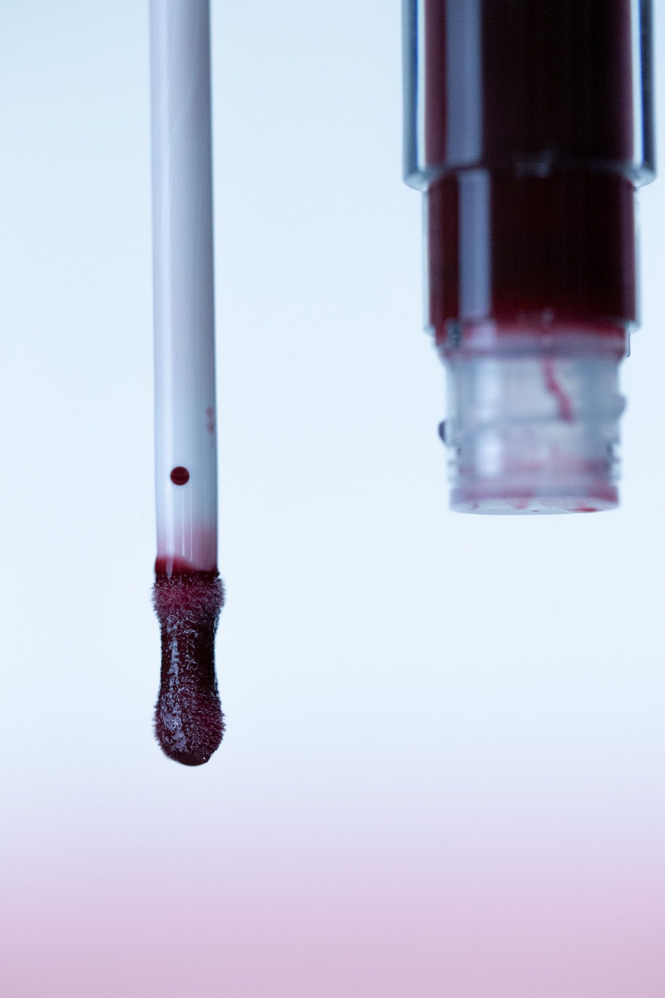Dark burgundy liquid lipstick applicator wand with product dripping on light gradient background.