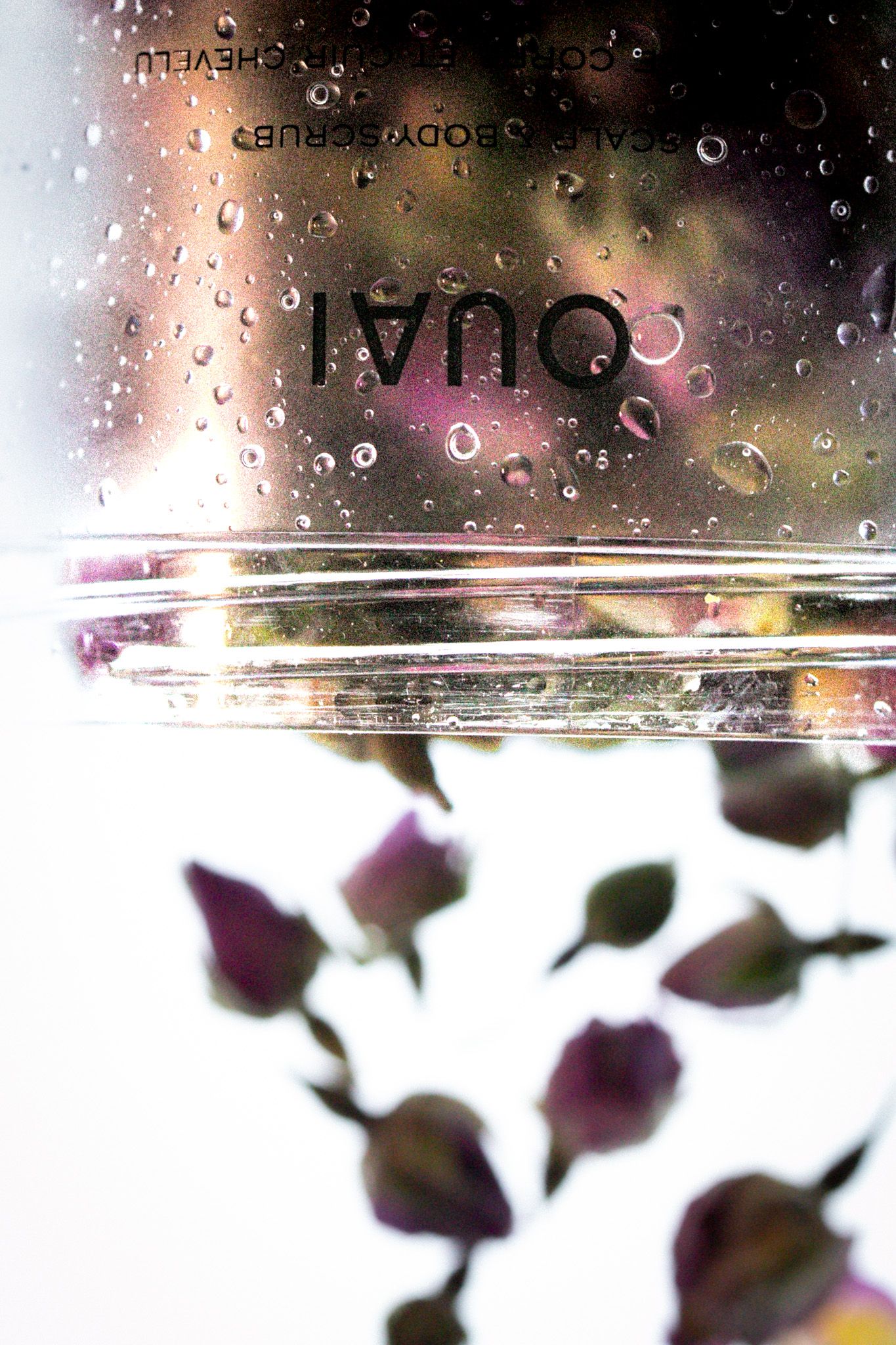 OUAI scalp and body scrub bottle with water droplets, surrounded by blurred dried rosebuds on white background.