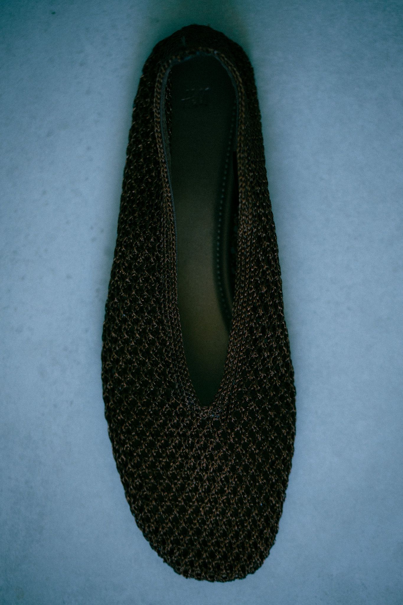 Black woven knit ballet flat shoe displayed on a blue surface, top-down view.