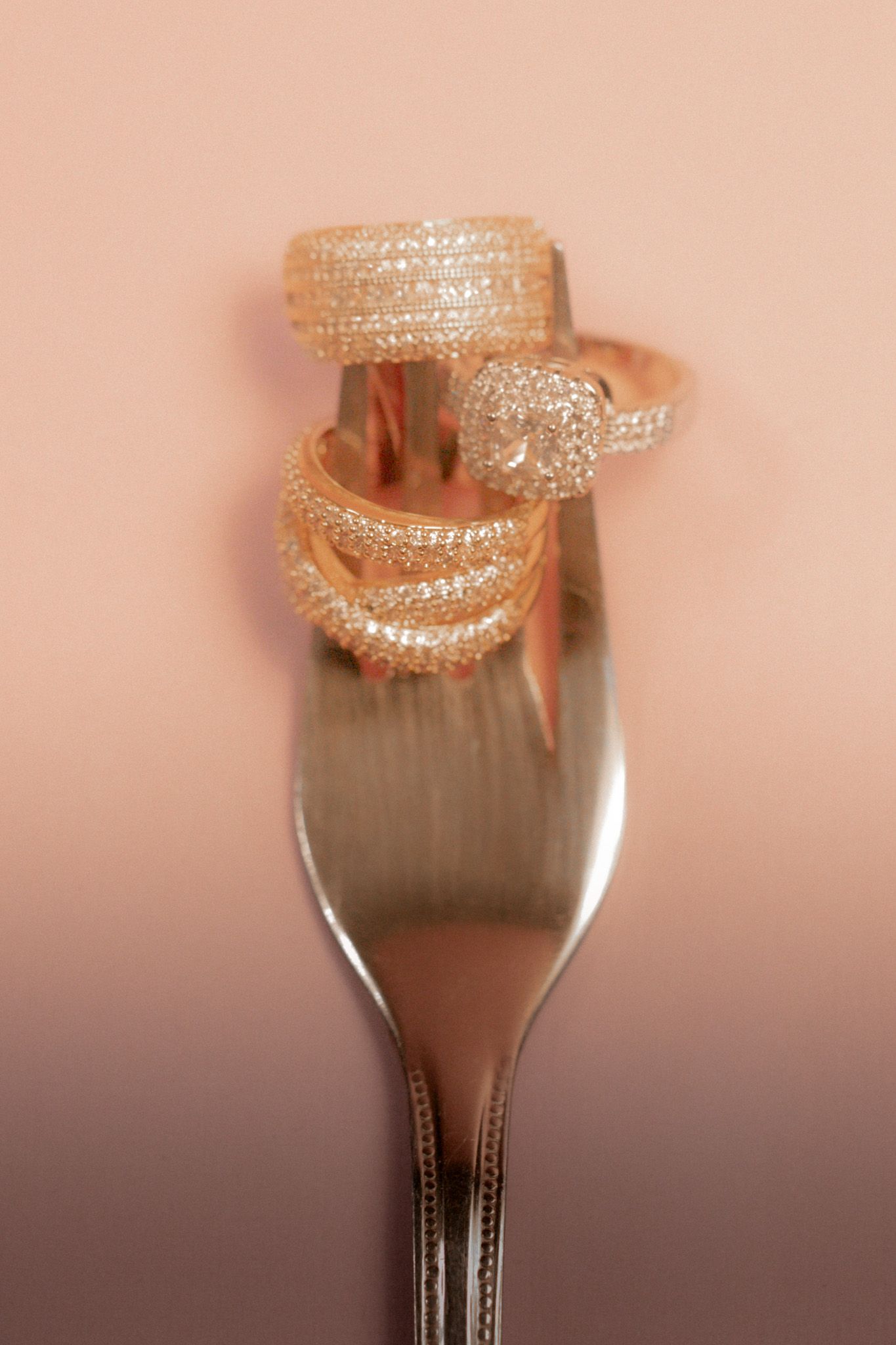 Three sparkling rose gold diamond rings stacked on silver fork tines on pink background.
