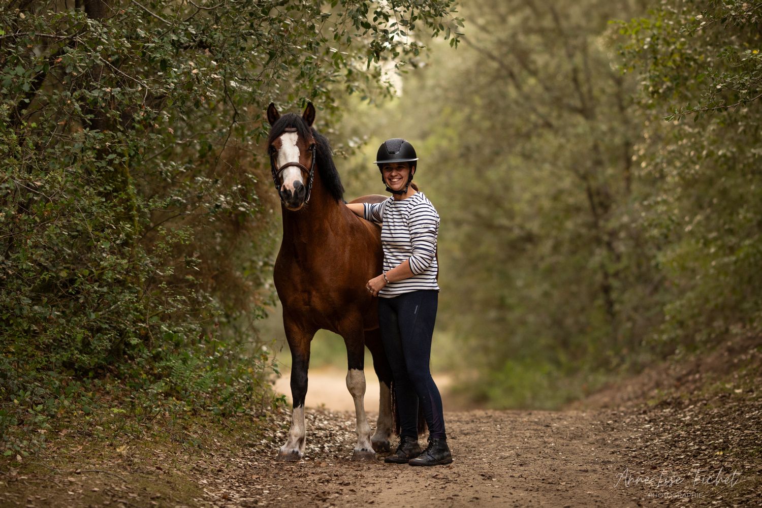 Rider in a helmet and striped shirt stands beside a brown horse with a white blaze on a shaded forest trail.