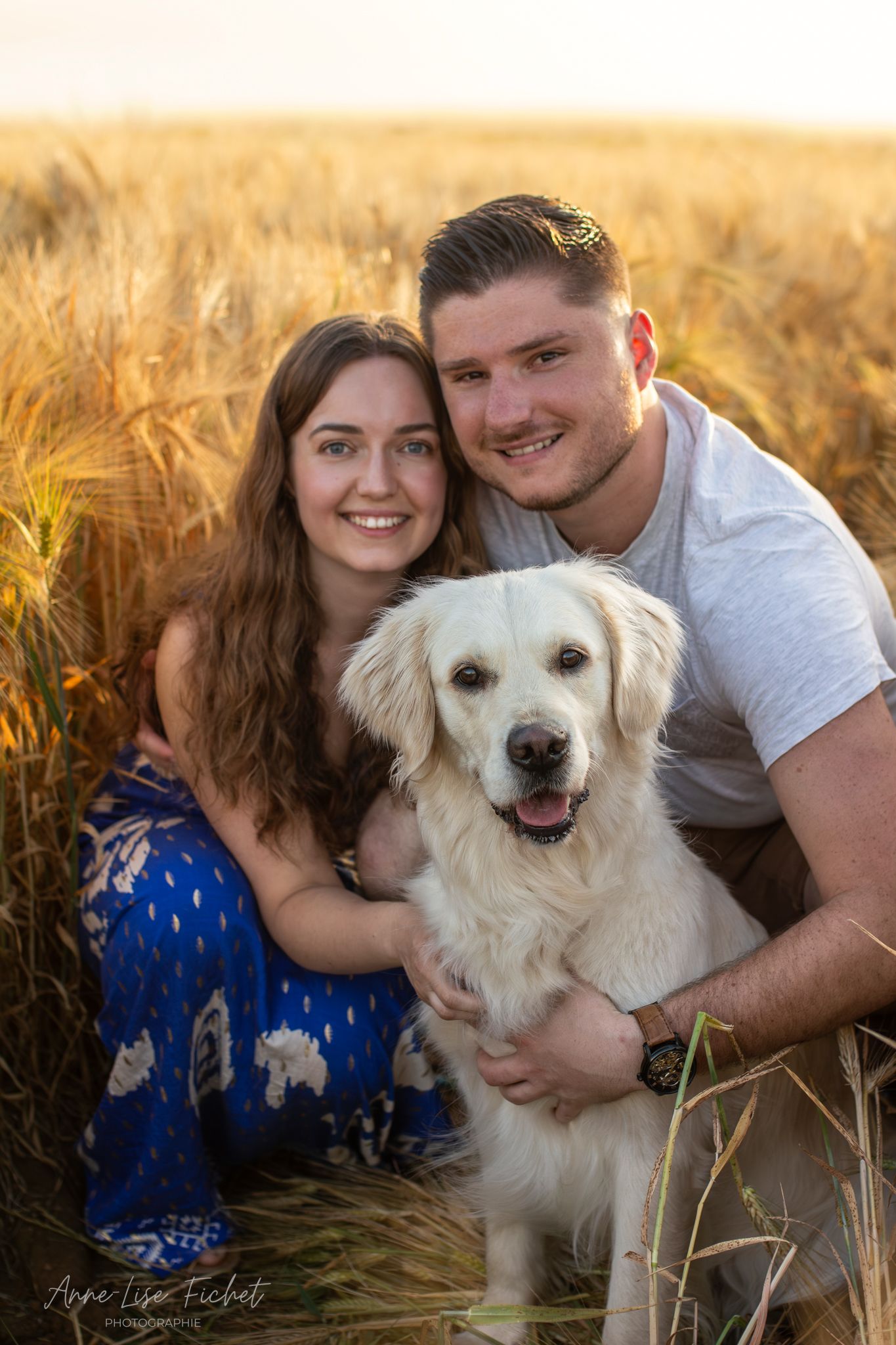 Smiling couple poses with a white Golden Retriever in a golden wheat field at sunset.